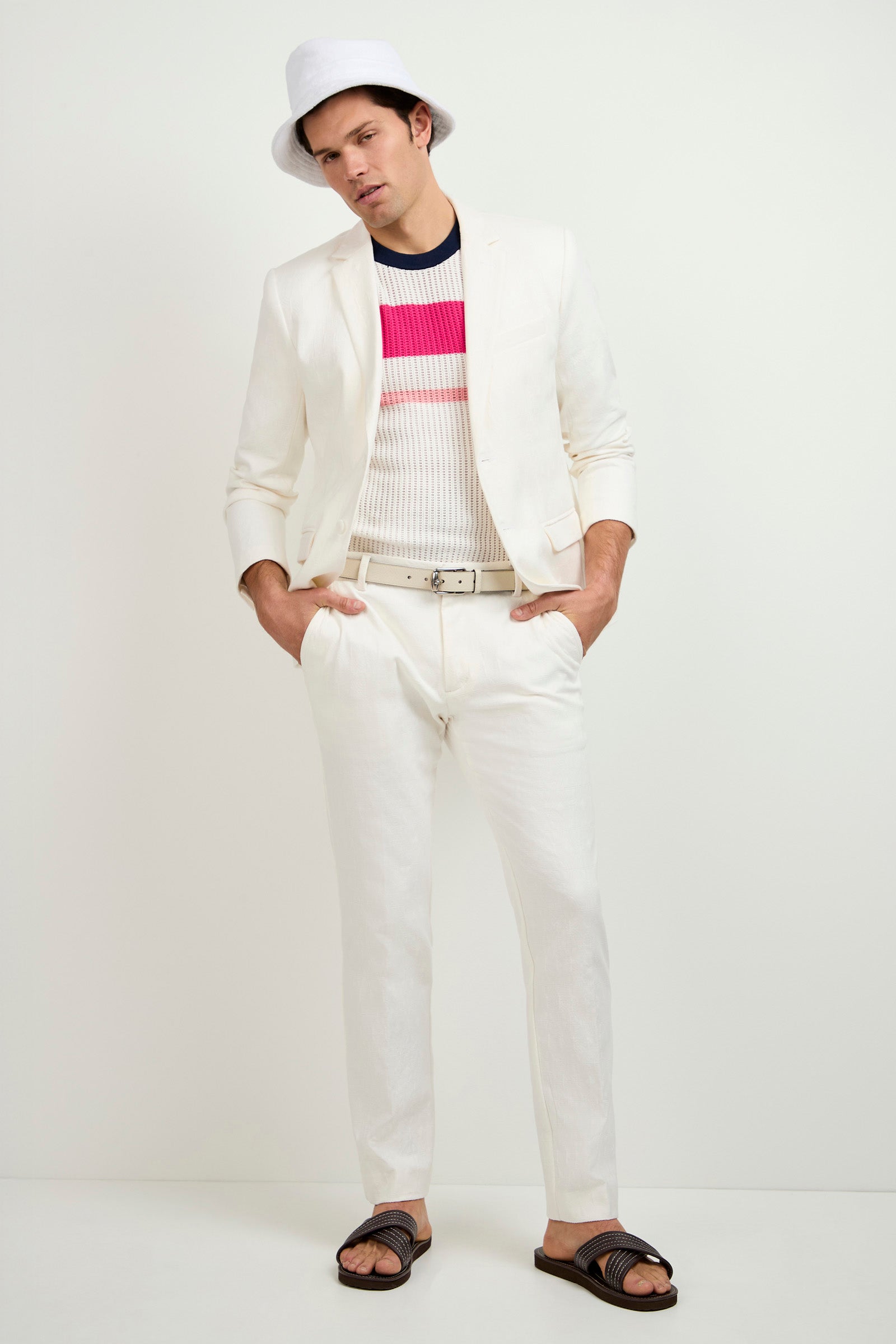 A man poses against a plain background wearing the Clyde Slim Trouser - Tegola Textured Jacquard paired with a white suit, white hat, ribbed striped sweater, white belt, and dark slide sandals. His hands are in his pockets as he faces the camera.