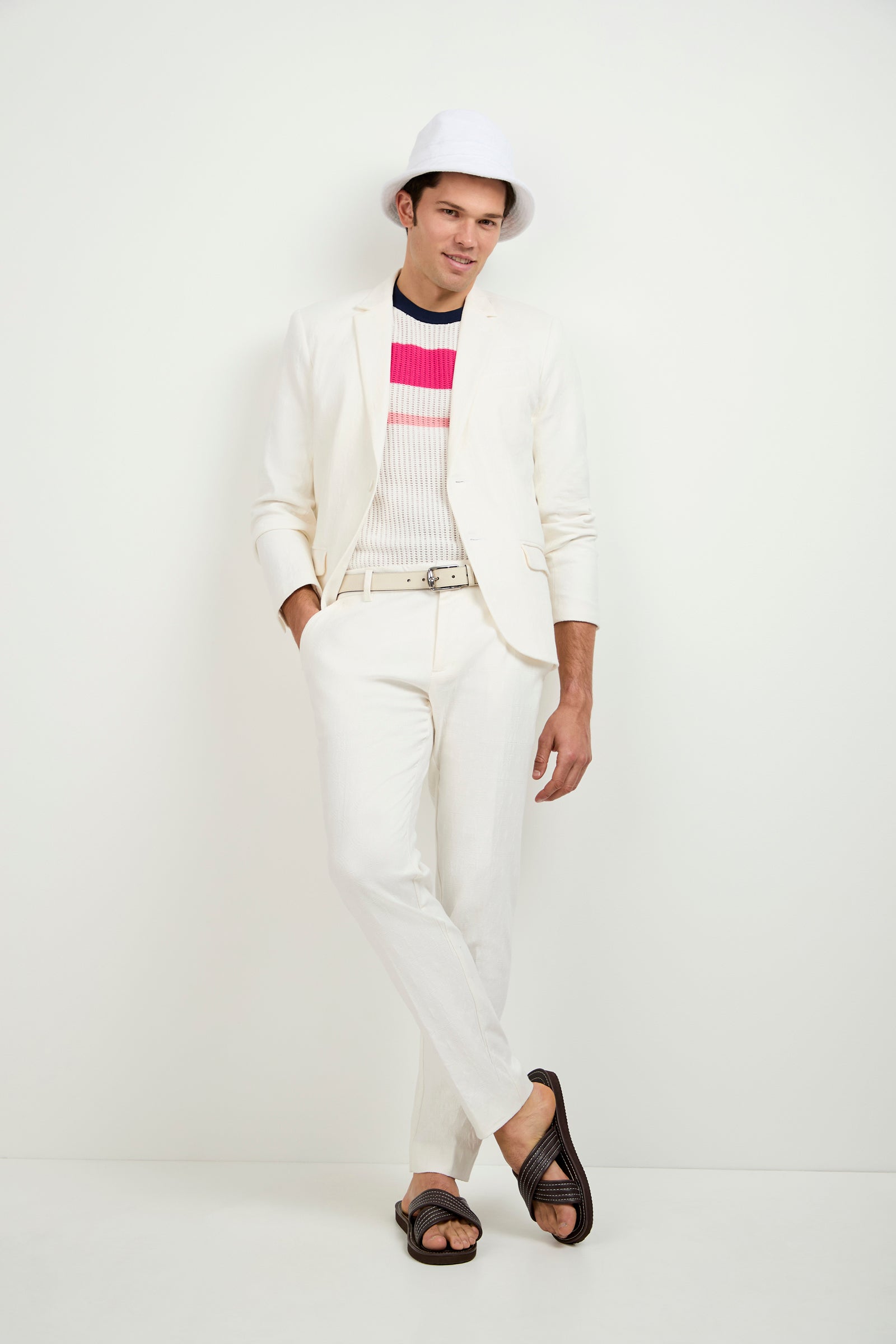 A man wearing the Thurston Blazer in Tegola Textured Jacquard, paired with a white bucket hat, striped pink and black sweater, and dark sandals stands against a plain white background with his legs crossed and one hand in his pocket.