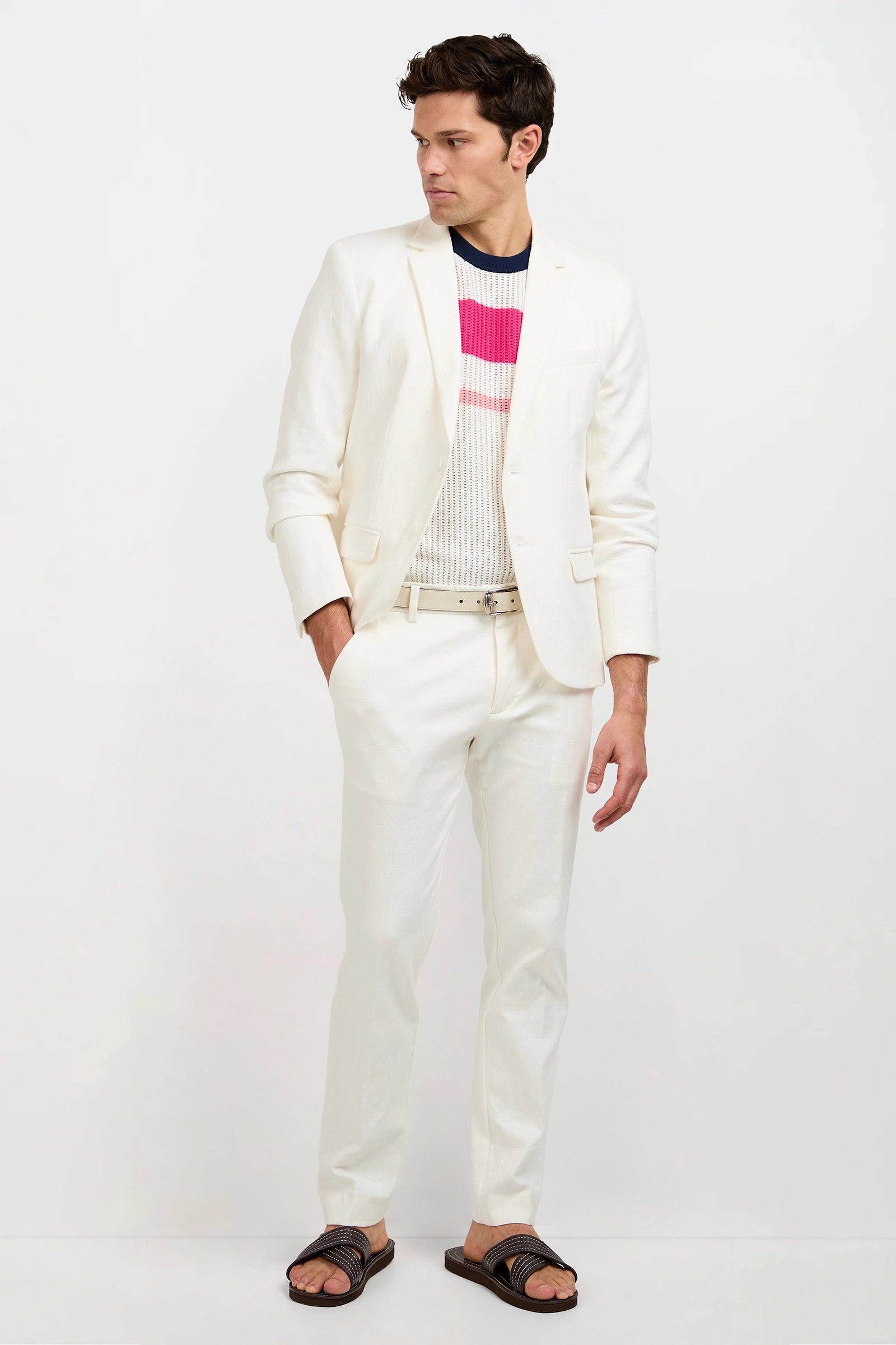 A man wearing the Thurston Blazer in Tegola Textured Jacquard, layered over a colorful striped sweater with dark sandals, stands against a plain white background, looking left with his right hand in his pocket.