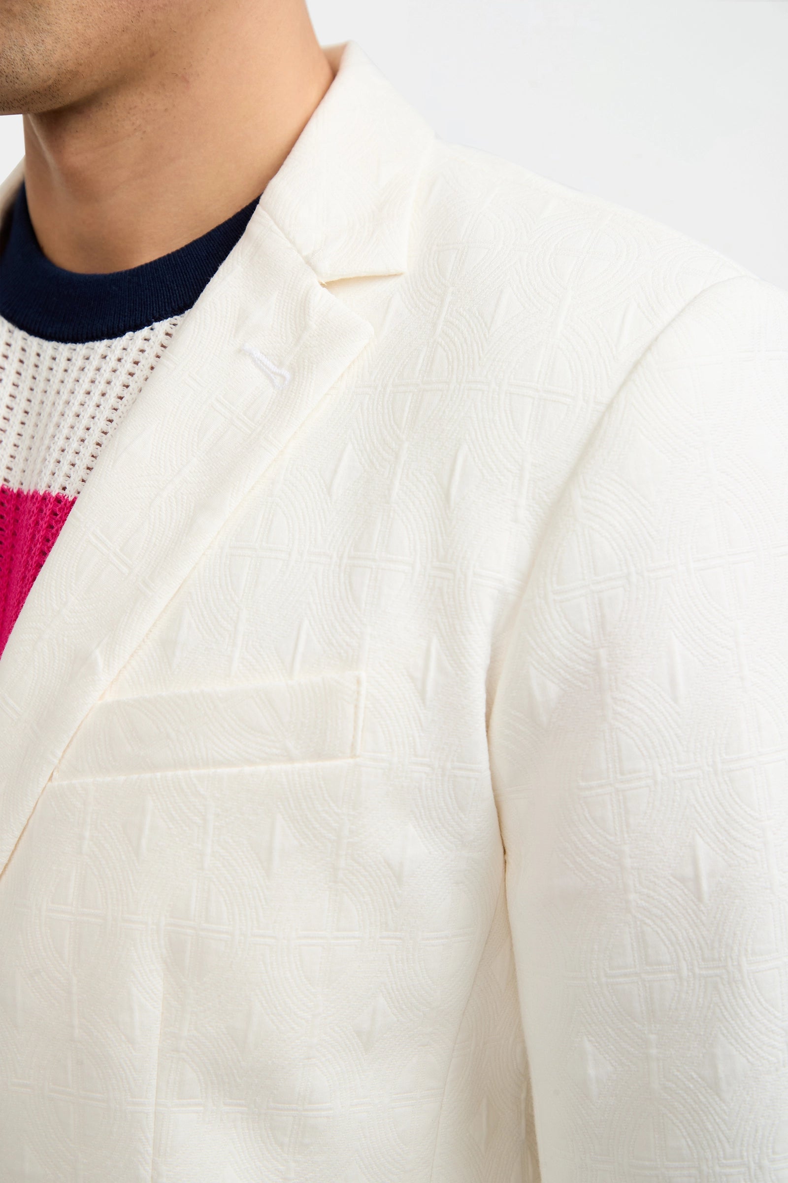 Close-up of a person wearing the Thurston Blazer - Tegola Textured Jacquard over a navy, white, and bright pink knit sweater. Only the upper chest and shoulder are visible against a plain light background.