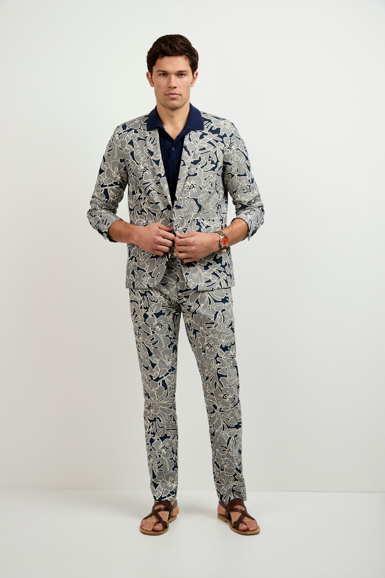 A man stands against a plain white background wearing the Thurston Blazer - Artsy Floral Jacquard over a dark blue shirt, paired with brown sandals and a watch, his hands fastening the blazer.