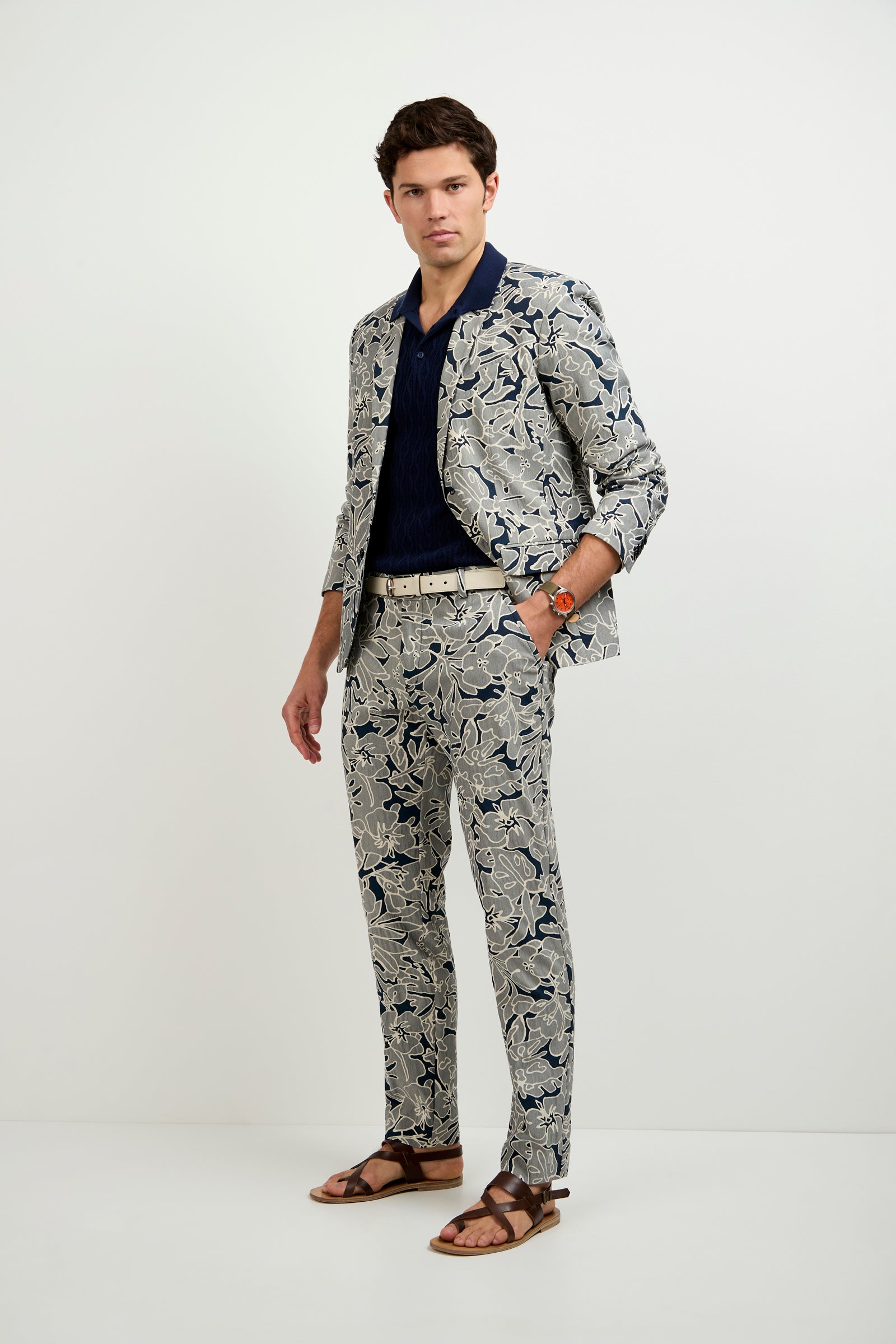 A man stands against a plain white background wearing the Thurston Blazer - Artsy Floral Jacquard, paired with a dark navy shirt, beige belt, brown sandals, an orange watch, and has one hand in his pocket.
