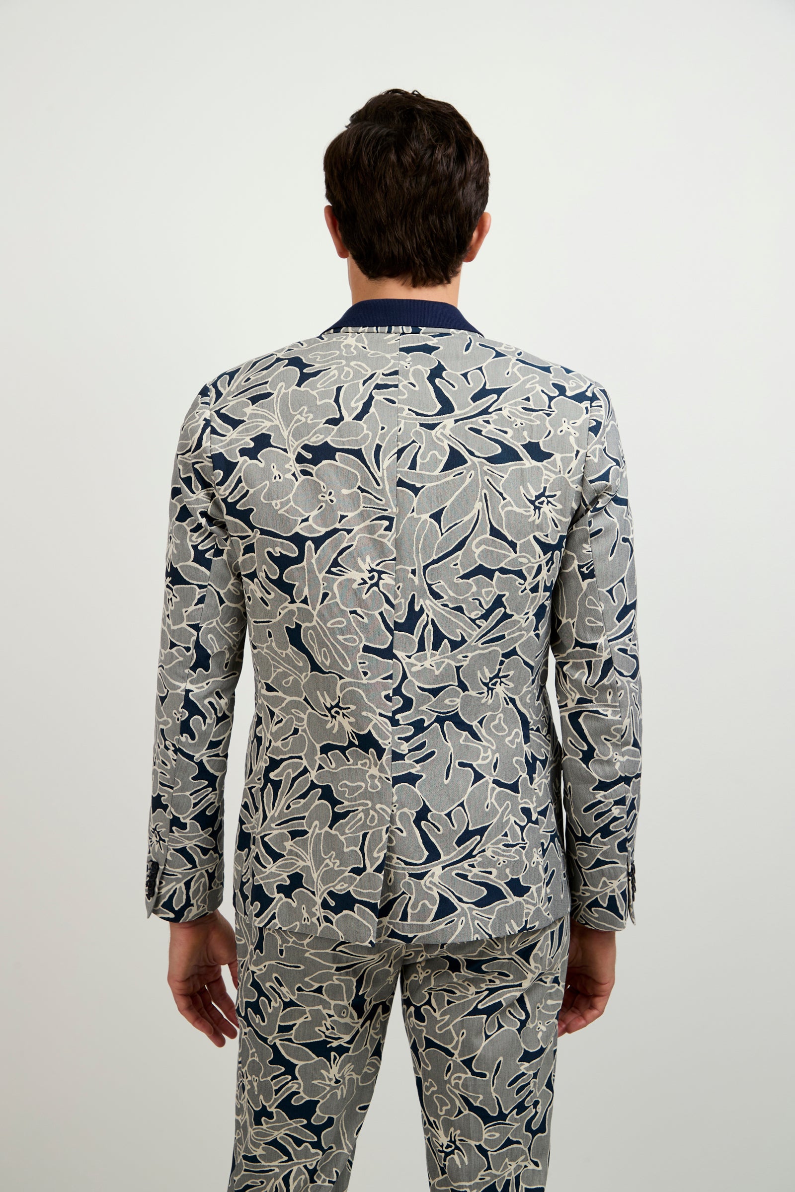 A person stands facing away, wearing the Thurston Blazer - Artsy Floral Jacquard, featuring a bold floral pattern in blue, gray, and beige against a plain light background.