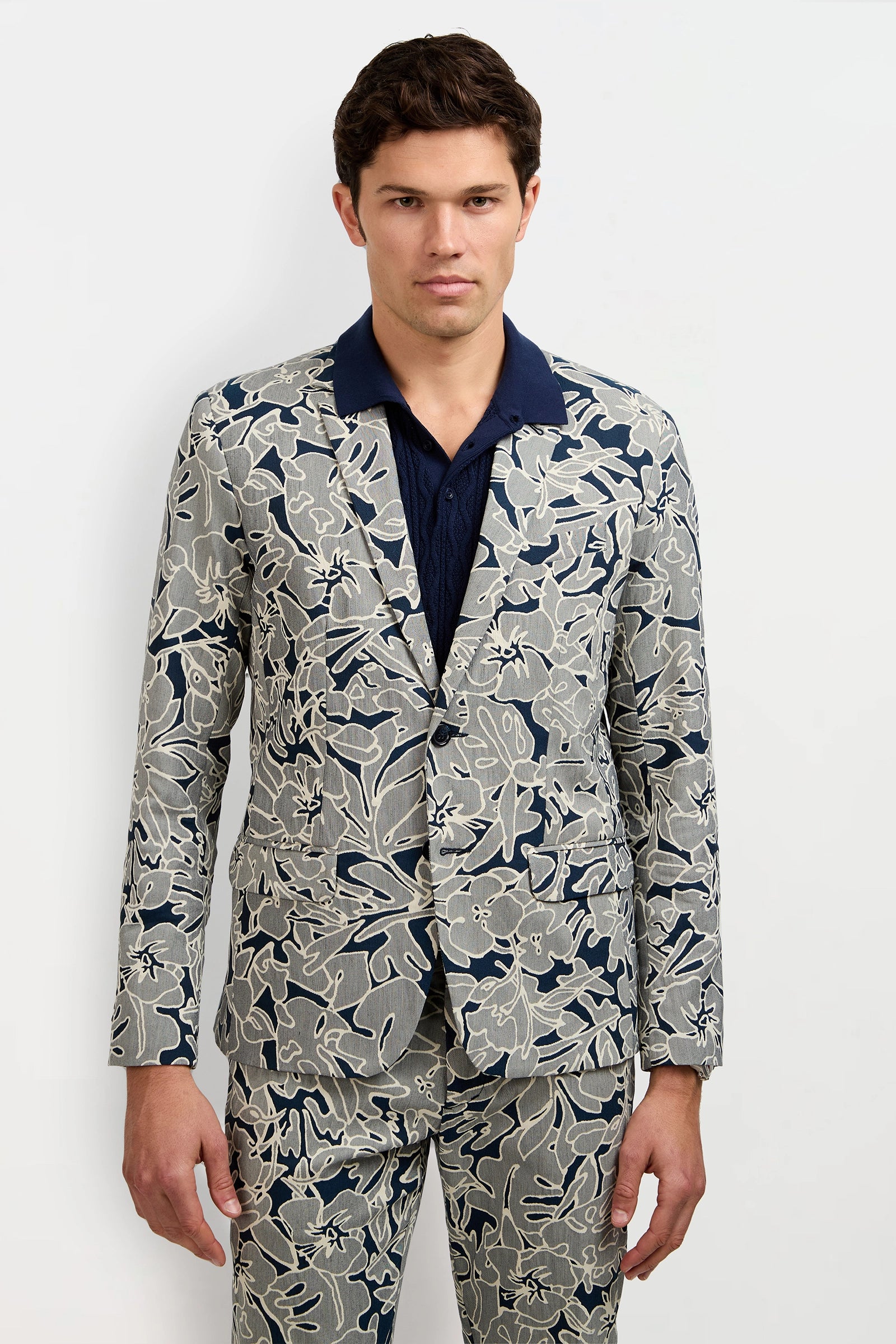A man with short dark hair and a neutral expression wears the Thurston Blazer - Artsy Floral Jacquard and matching pants, featuring a bold blue and white floral pattern, over a dark navy collared shirt against a plain background.