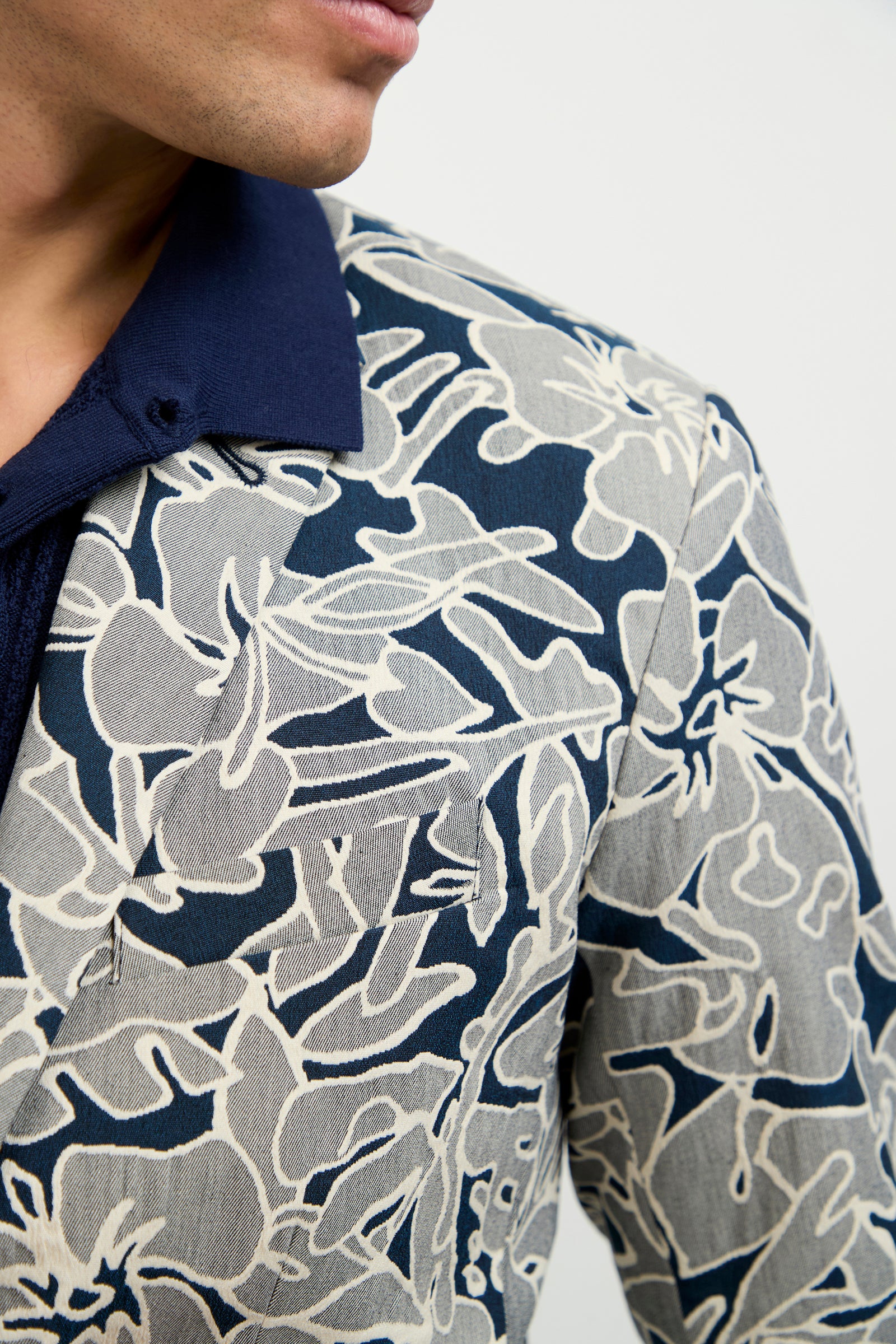 A close-up shows a person in the Thurston Blazer - Artsy Floral Jacquard, featuring a bold beige and navy abstract floral pattern over a navy shirt. Only the lower face and shoulder are seen against a plain background.
