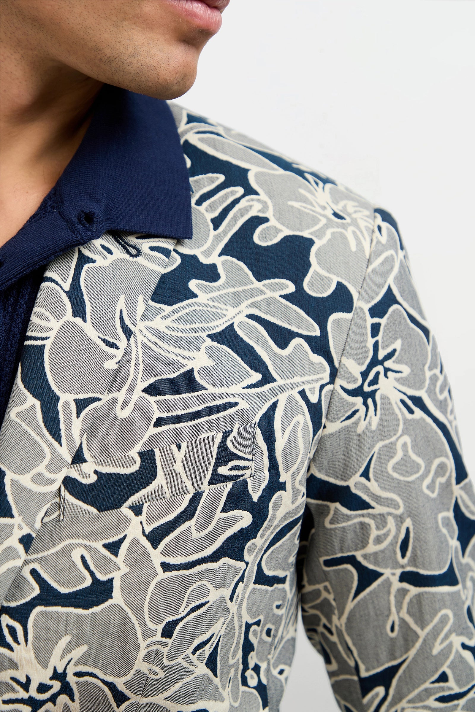 A person wears the Thurston Blazer - Artsy Floral Jacquard, featuring a bold cream floral pattern over a navy shirt. The person's face is partly visible against a plain white background.