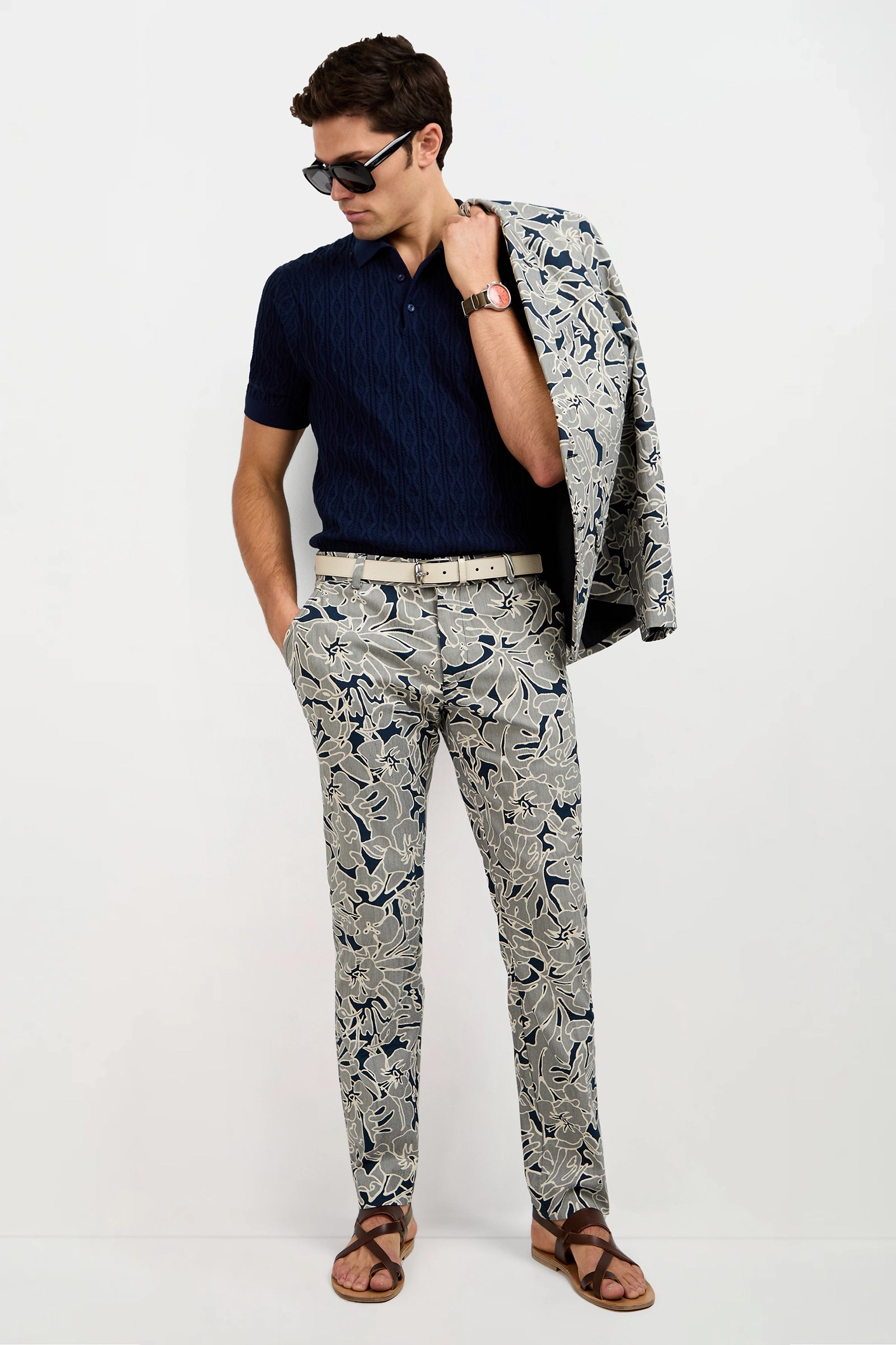 A man stands against a plain white background, hands in pockets, wearing sunglasses, a navy polo shirt, and Clyde Slim Trouser - Artsy Floral Jacquard with the matching blazer draped over his shoulder.