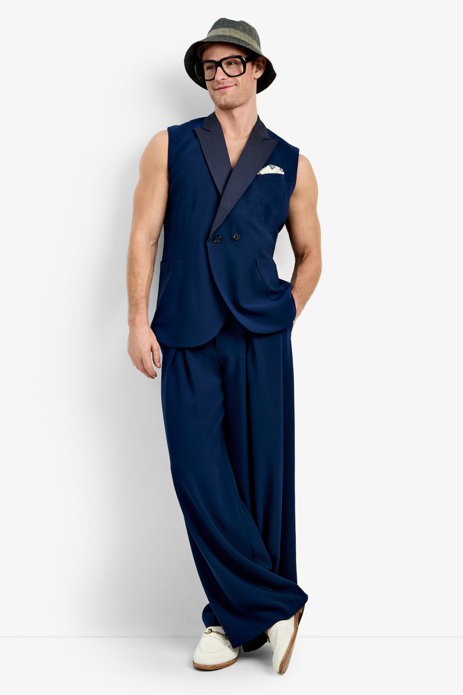 A man stands against a white background, smiling with one hand in his pocket, wearing the Thurman Sleeveless Blazer, navy wide-leg pants, a gray hat, black glasses, a white pocket square, and white shoes.