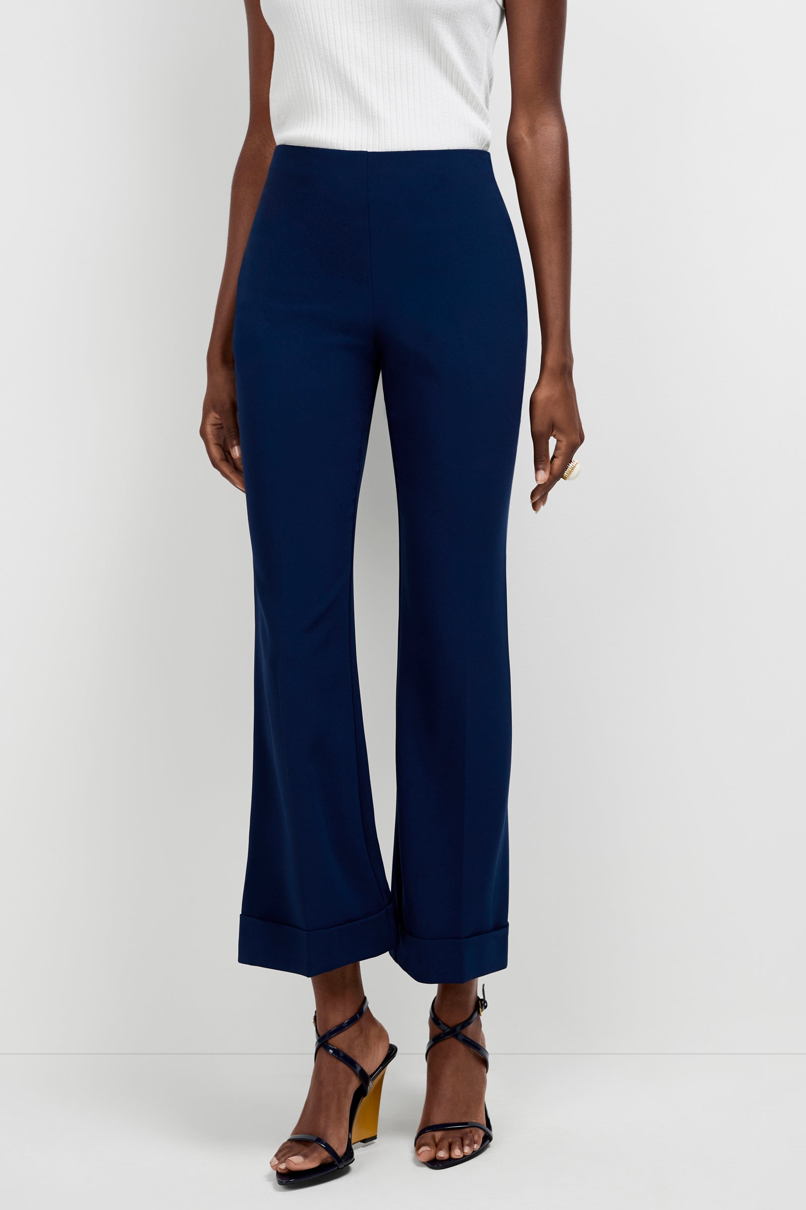 A person wearing a sleeveless white top and Loren Cropped Kick-Flare Pants in dark blue with cuffed hems, paired with black strappy heeled sandals, stands against a plain white background.