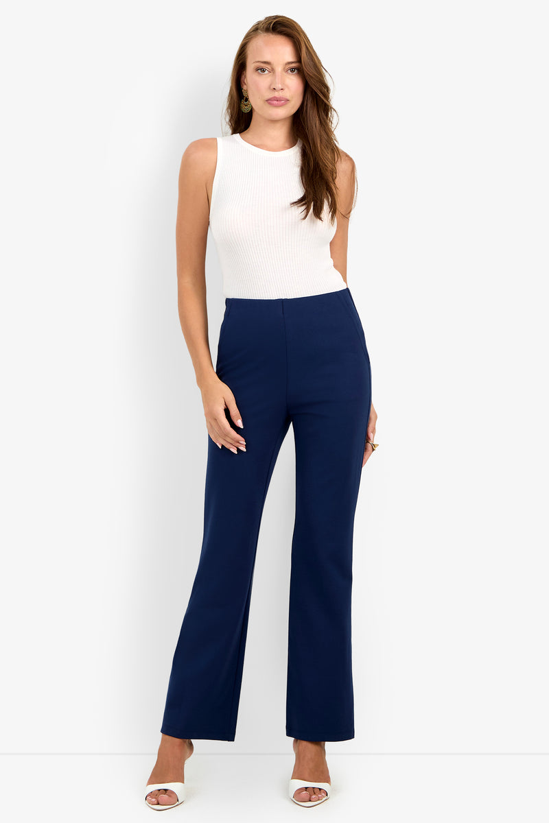 A woman stands against a white background wearing a sleeveless white top, the Luisa Pant in navy blue, and white heeled sandals.
