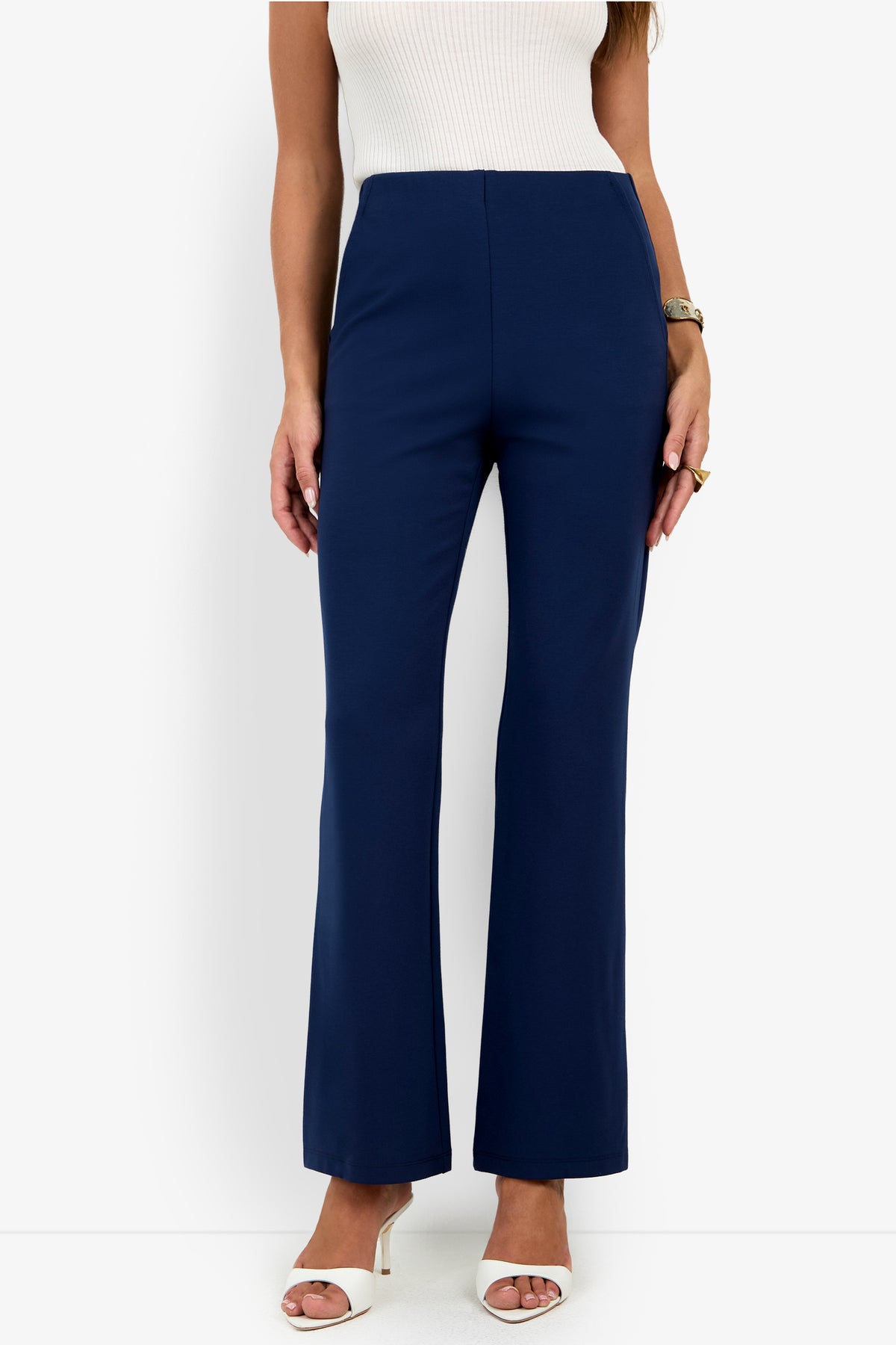 A woman wearing a sleeveless white top, the Luisa Pant in navy blue, white heeled sandals, a watch, and rings stands against a plain white background.