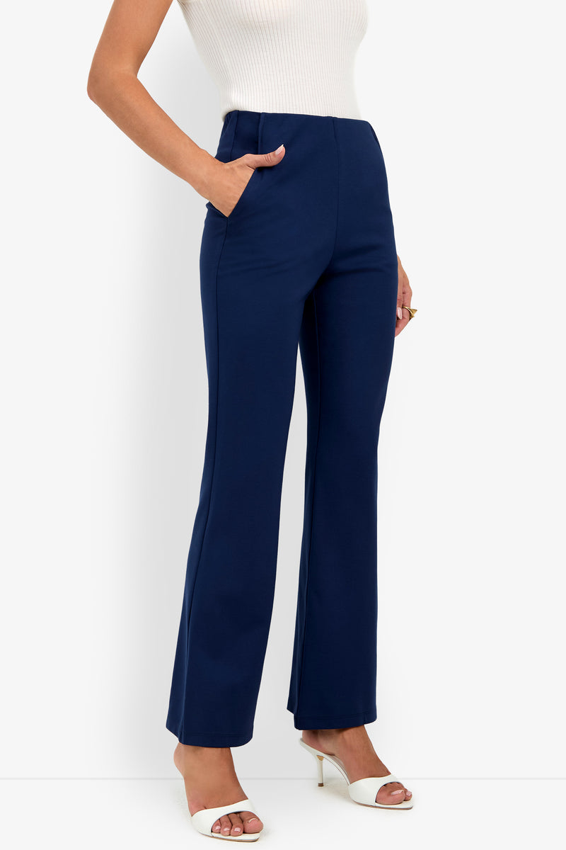 A person wearing the Luisa Pant, a high-waisted, navy blue flare style, paired with white heeled sandals and one hand in a pocket, stands against a plain white background.