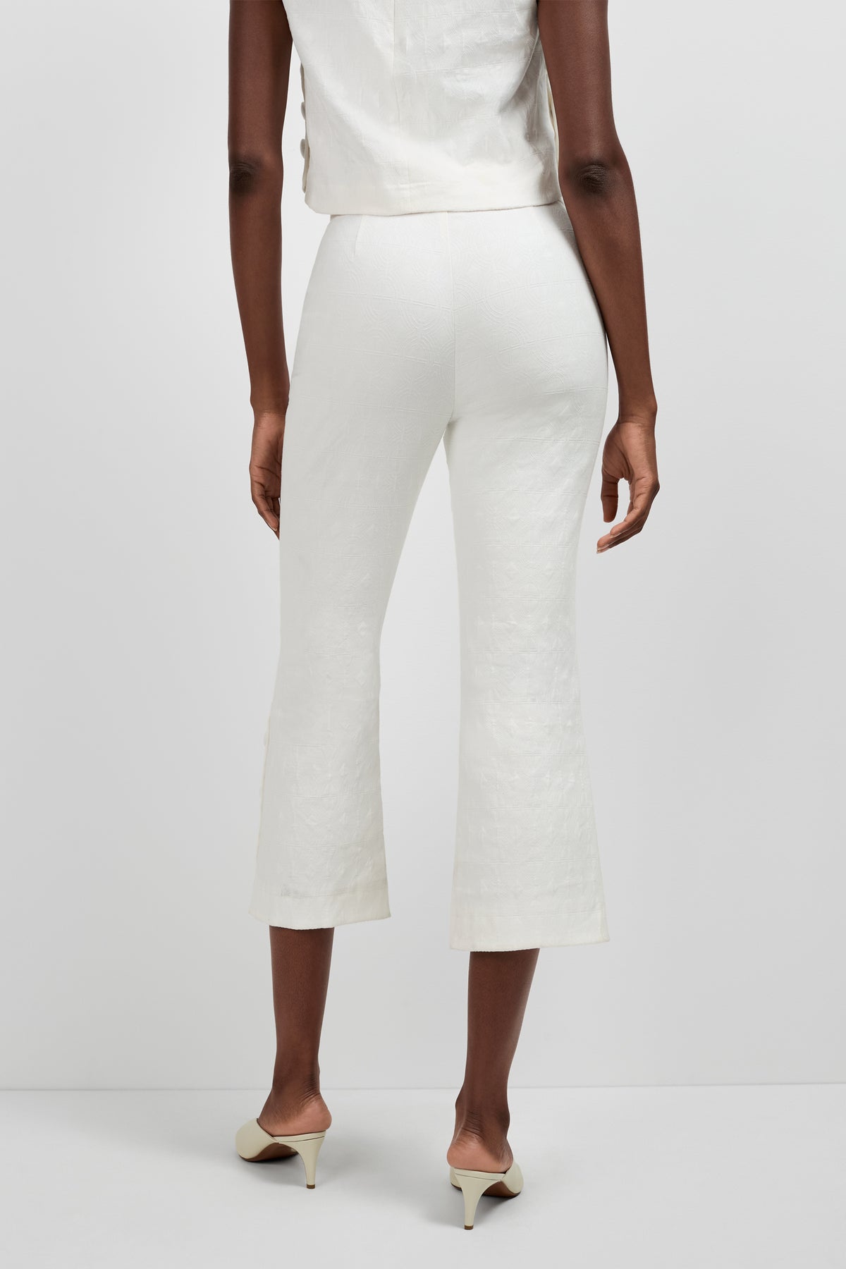 A person stands facing away, wearing Tortona Button Hem Pants in off-white, a matching sleeveless top, and cream-colored heeled shoes against a plain light background.