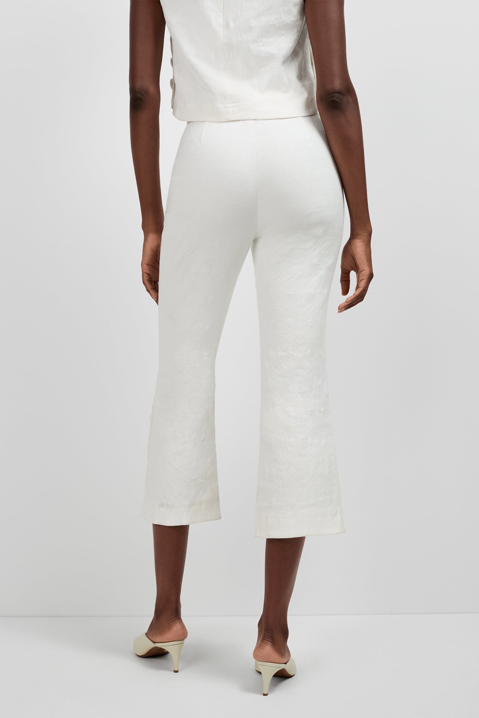 A person stands facing away, wearing Tortona Button Hem Pants in off-white, a matching sleeveless top, and cream-colored heeled shoes against a plain light background.