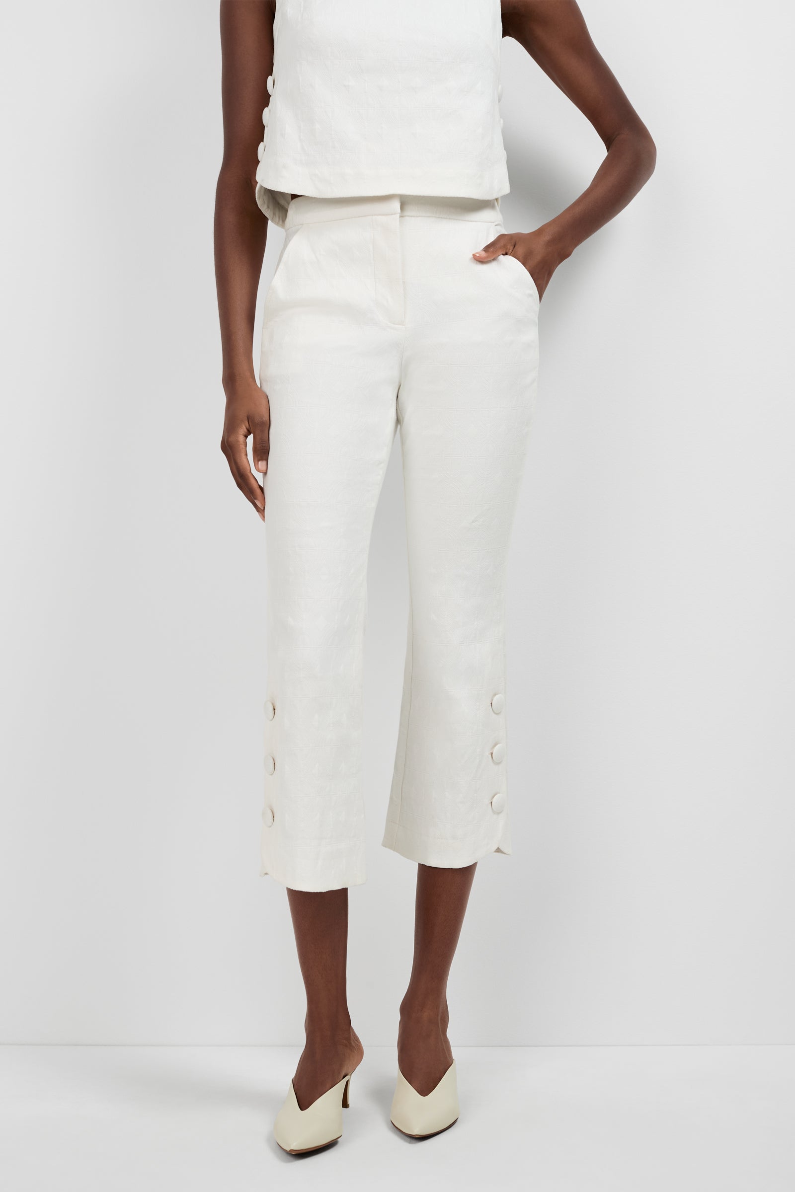 Someone stands against a plain white background wearing the Tortona Button Hem Pants—white cropped pants with buttons near the hem—a matching sleeveless top, and cream heeled shoes, with one hand in their pocket.
