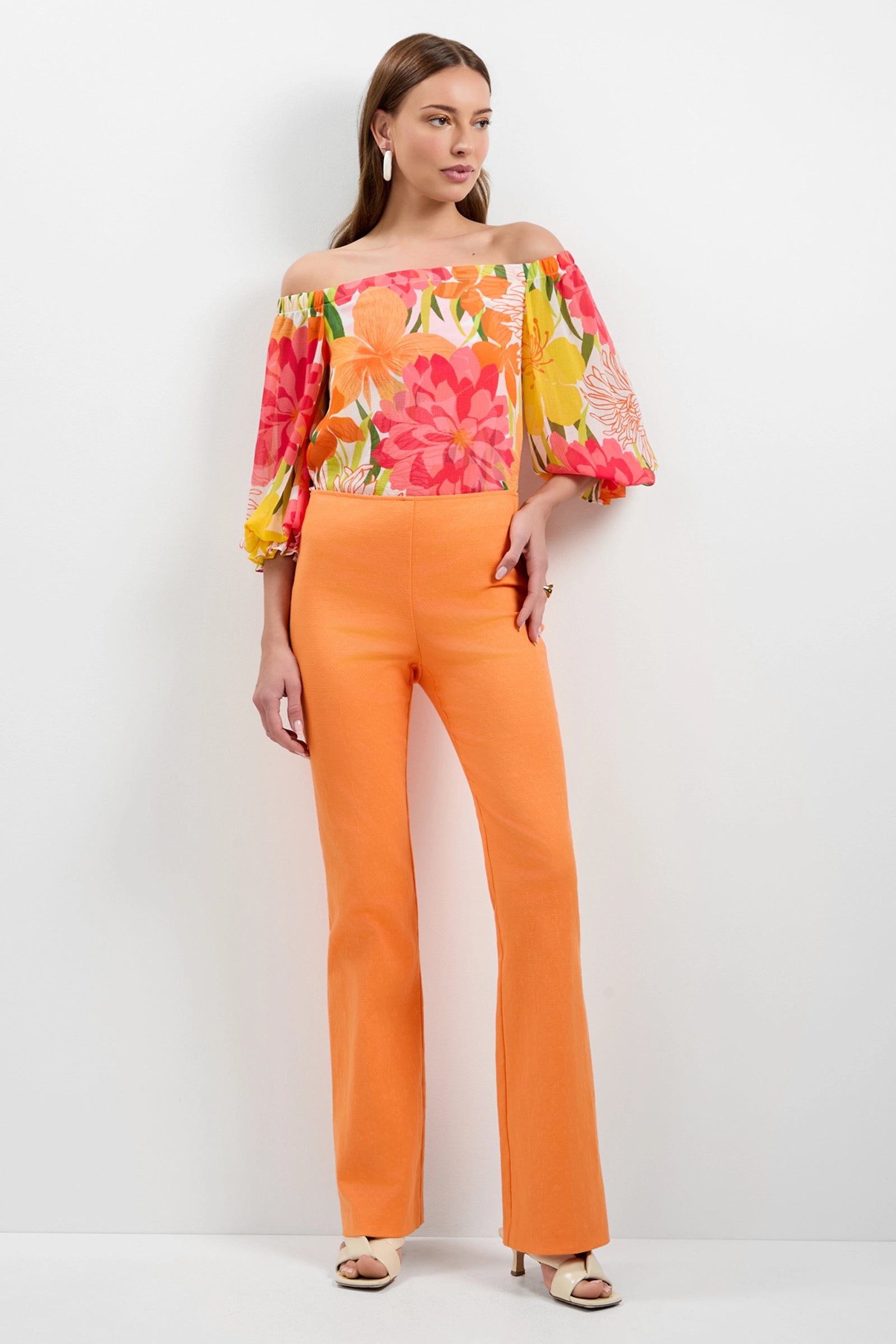 A woman stands against a white background wearing Jacoba Slim-Fit Bootcut Pants in orange and a colorful off-the-shoulder floral blouse with pink, orange, and yellow accents. She completes the look with beige open-toe heels.