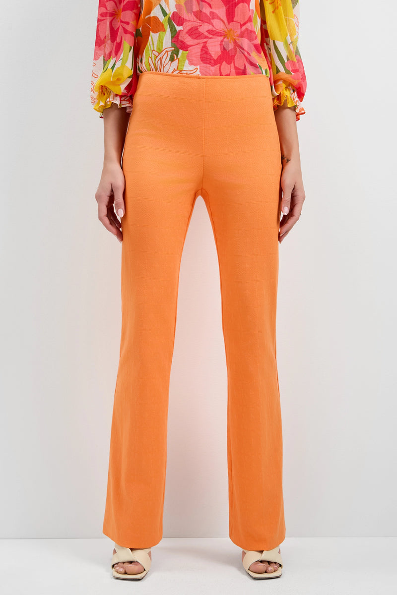 A person wears Jacoba Slim-Fit Bootcut Pants in bright orange with open-toed beige heels and a colorful floral long-sleeve top. The lower body is highlighted against a plain white background.