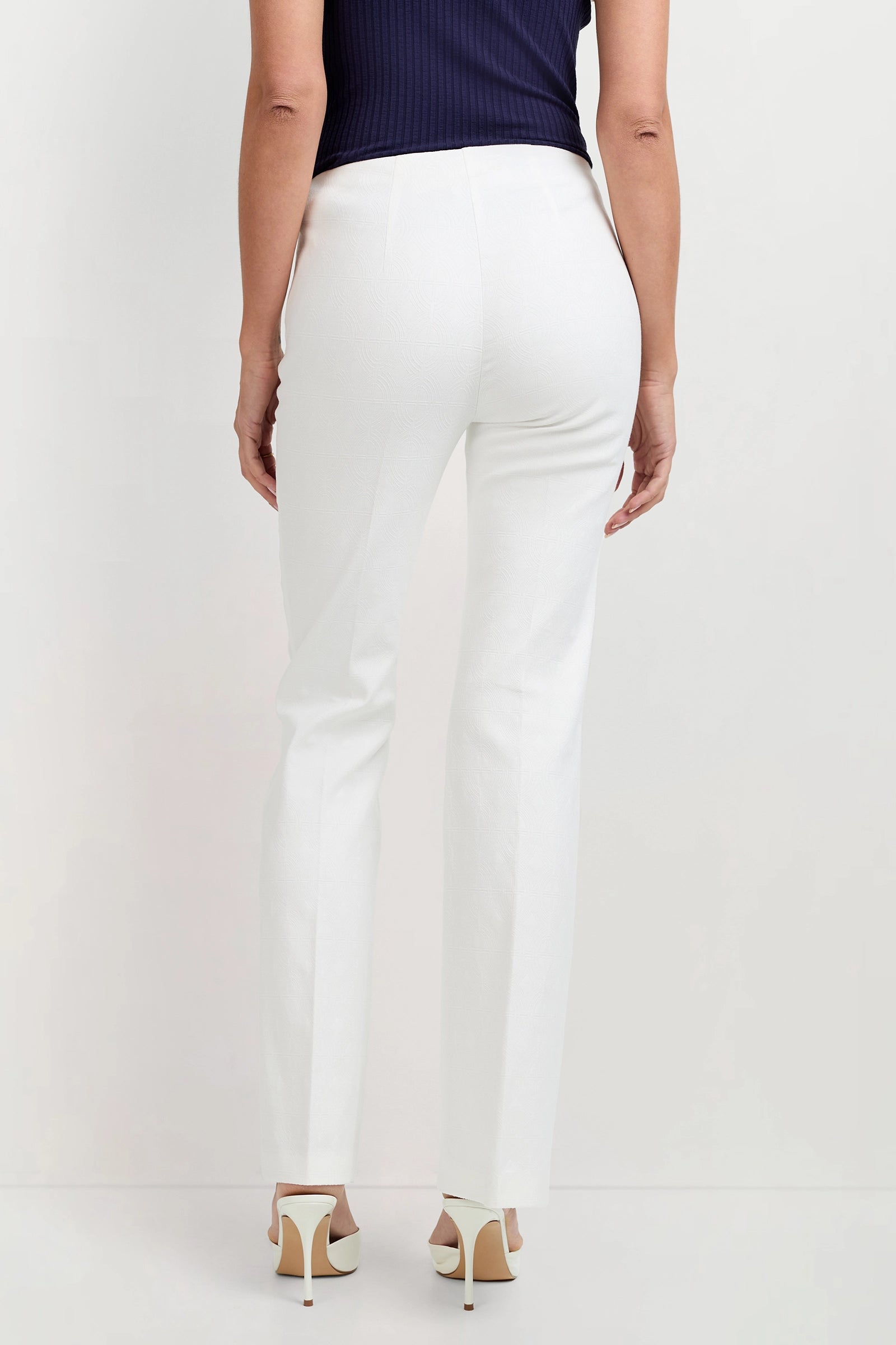 Someone stands with their back to the camera against a light background, wearing Jacoba Slim-Fit Bootcut Pants in white, a dark sleeveless top, and white high-heeled shoes.