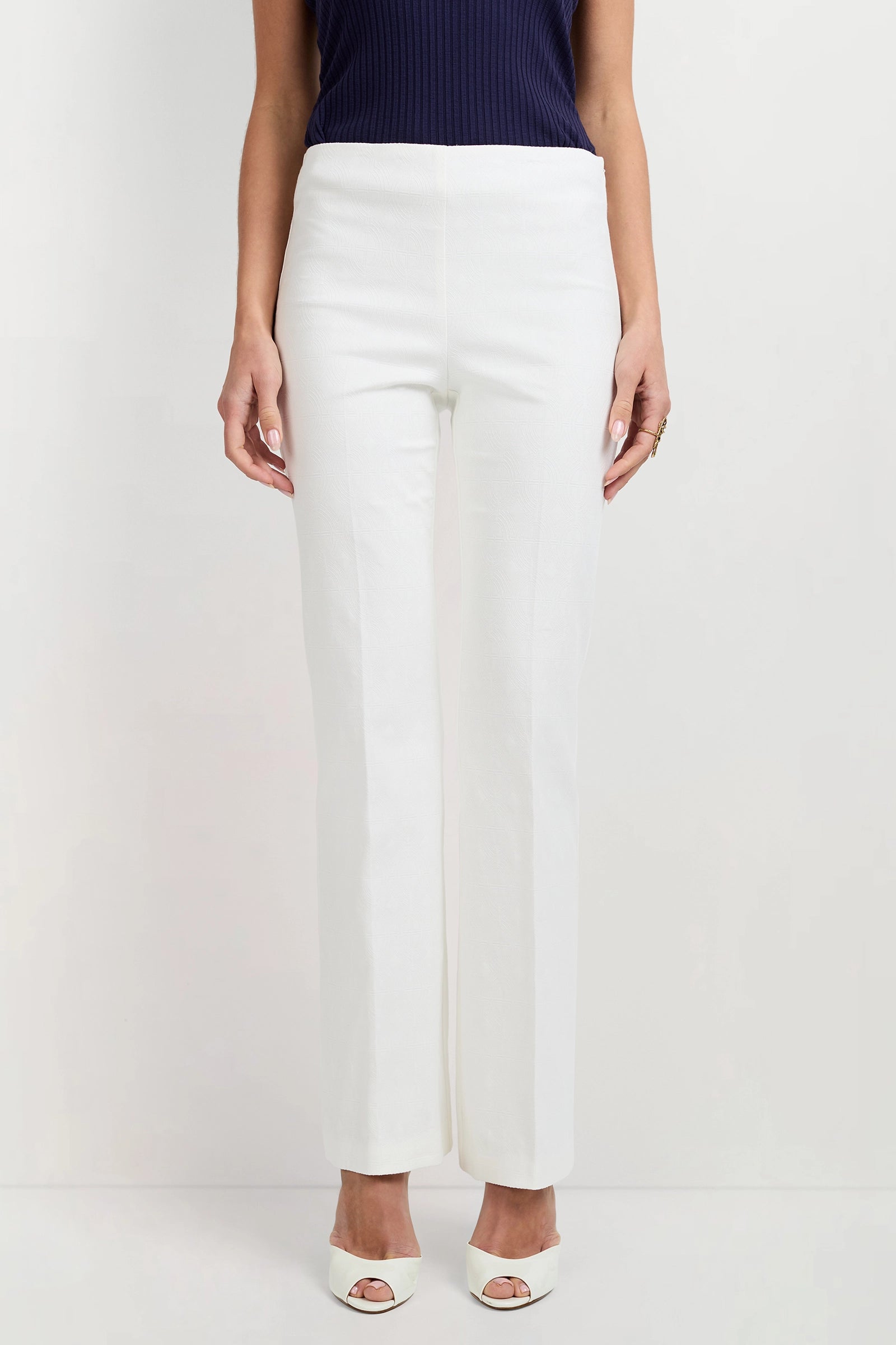 A person stands against a plain white background wearing Jacoba Slim-Fit Bootcut Pants in white, a sleeveless navy blue top, and white open-toe heels.
