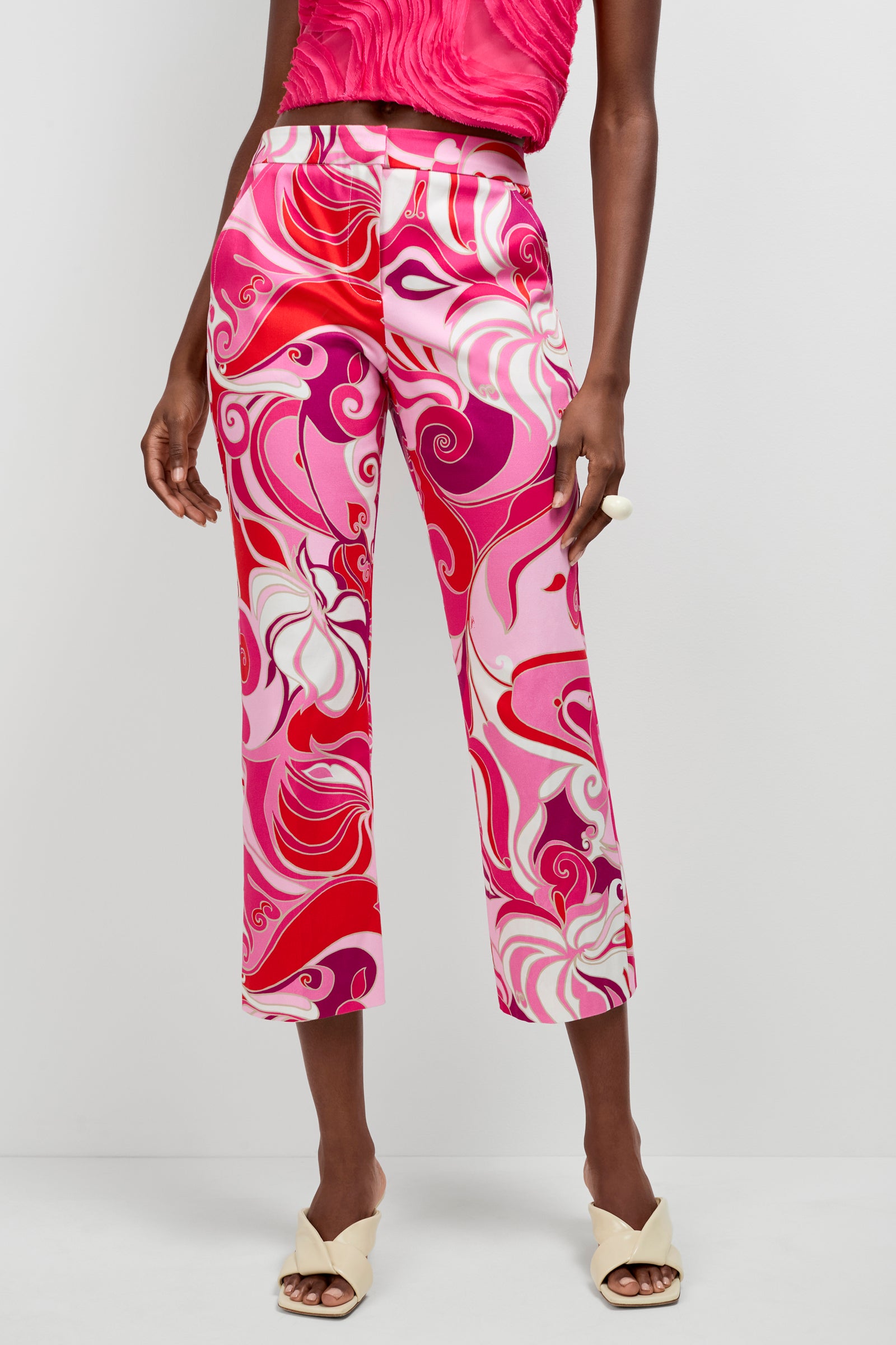 A person wears the Lulu Cropped Pants - Darsana, featuring a vibrant pink, red, and white swirling pattern, paired with a sleeveless pink top, cream open-toe heeled sandals, and a chunky white ring against a plain background.