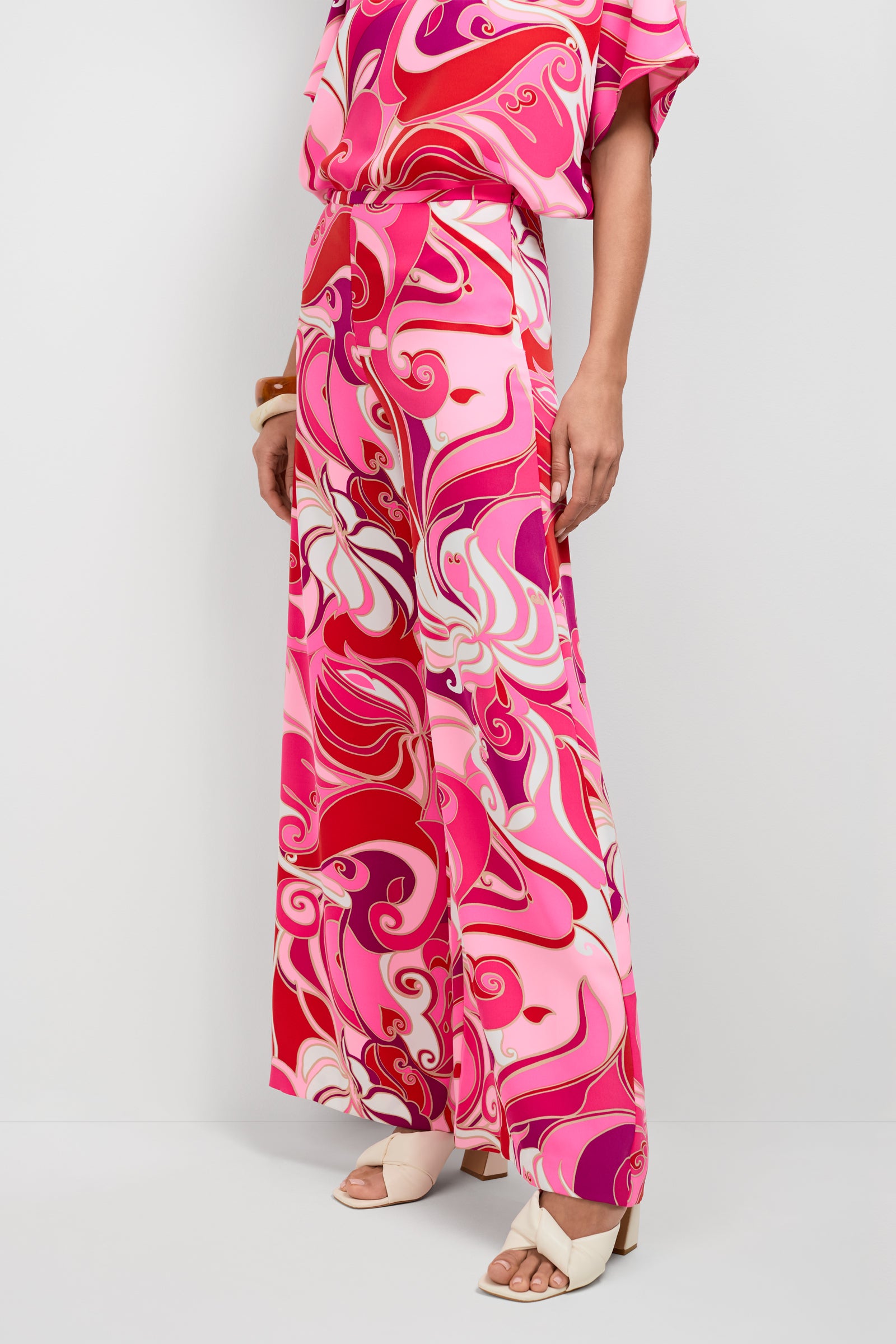 Someone stands against a plain white background wearing the Long Weekend Pants—wide-leg trousers with a bold, swirling pink, red, and white pattern—paired with a matching top and white open-toe heels.