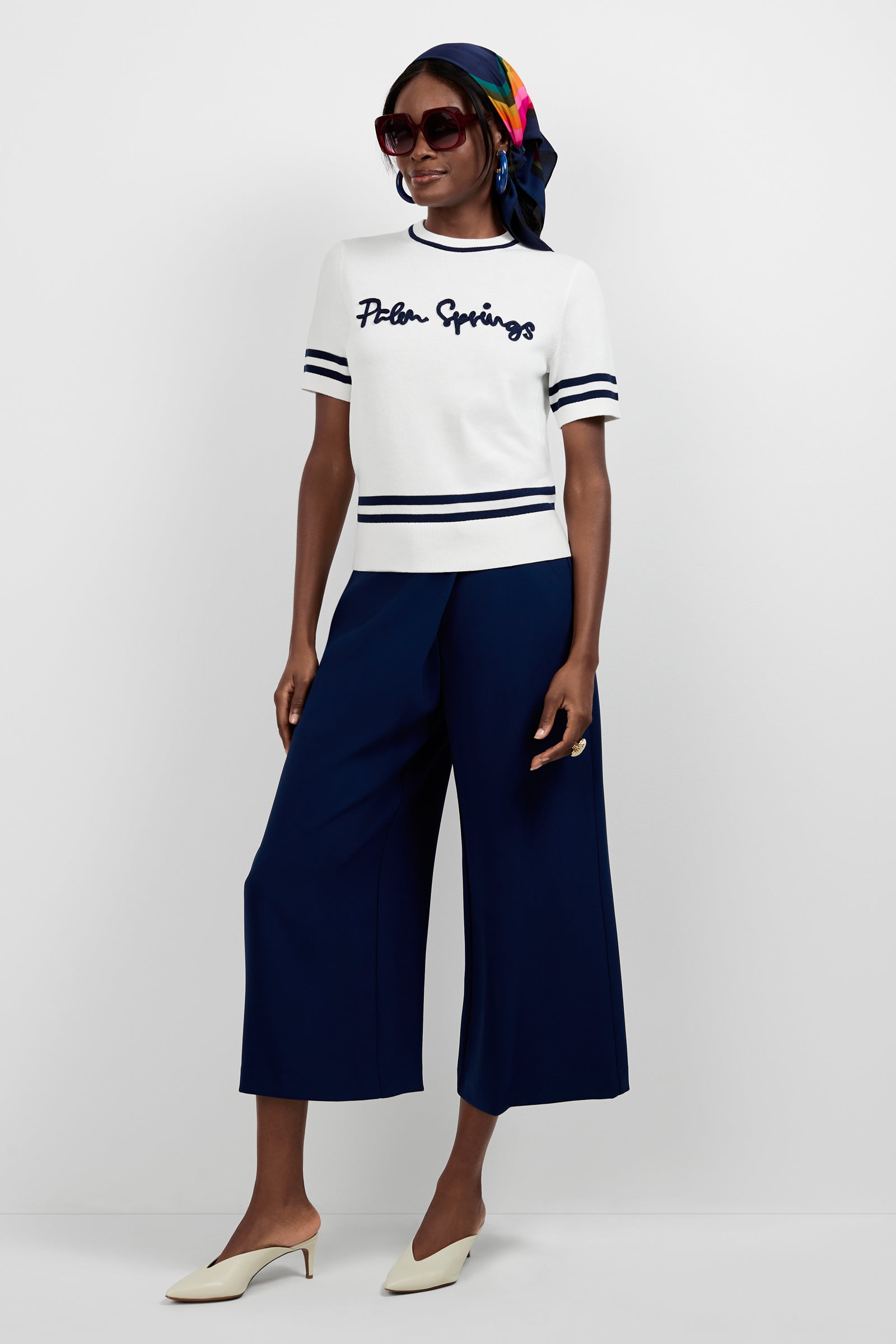 A woman smiles against a white background, wearing sunglasses, a colorful headscarf, a white Palm Springs striped short-sleeve sweater, Conchetta Wide-Leg Pants in navy, and cream heeled mules.