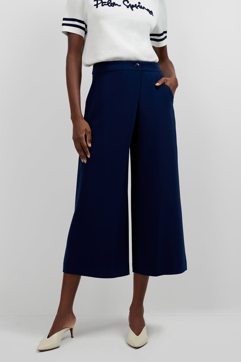 Wearing a white short-sleeve shirt with dark sleeve stripes, Conchetta Wide-Leg Pants in navy, and white heeled shoes, this person stands with one hand in their pocket.