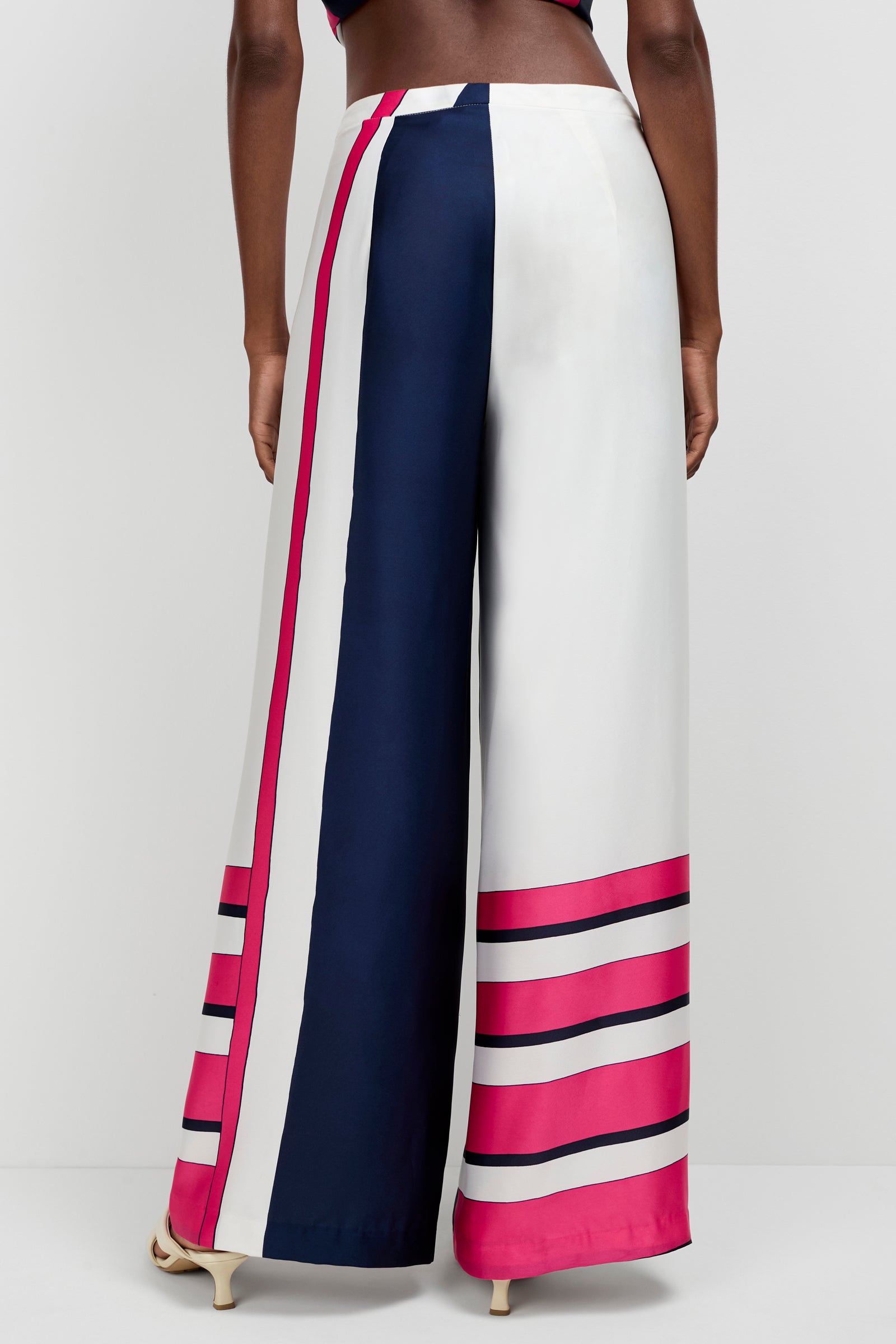 A person seen from the back wears the Lanay Linee Wide-Leg Panta, featuring vertical color-blocked stripes in navy, white, and magenta, with horizontal magenta and navy stripes near the hem.