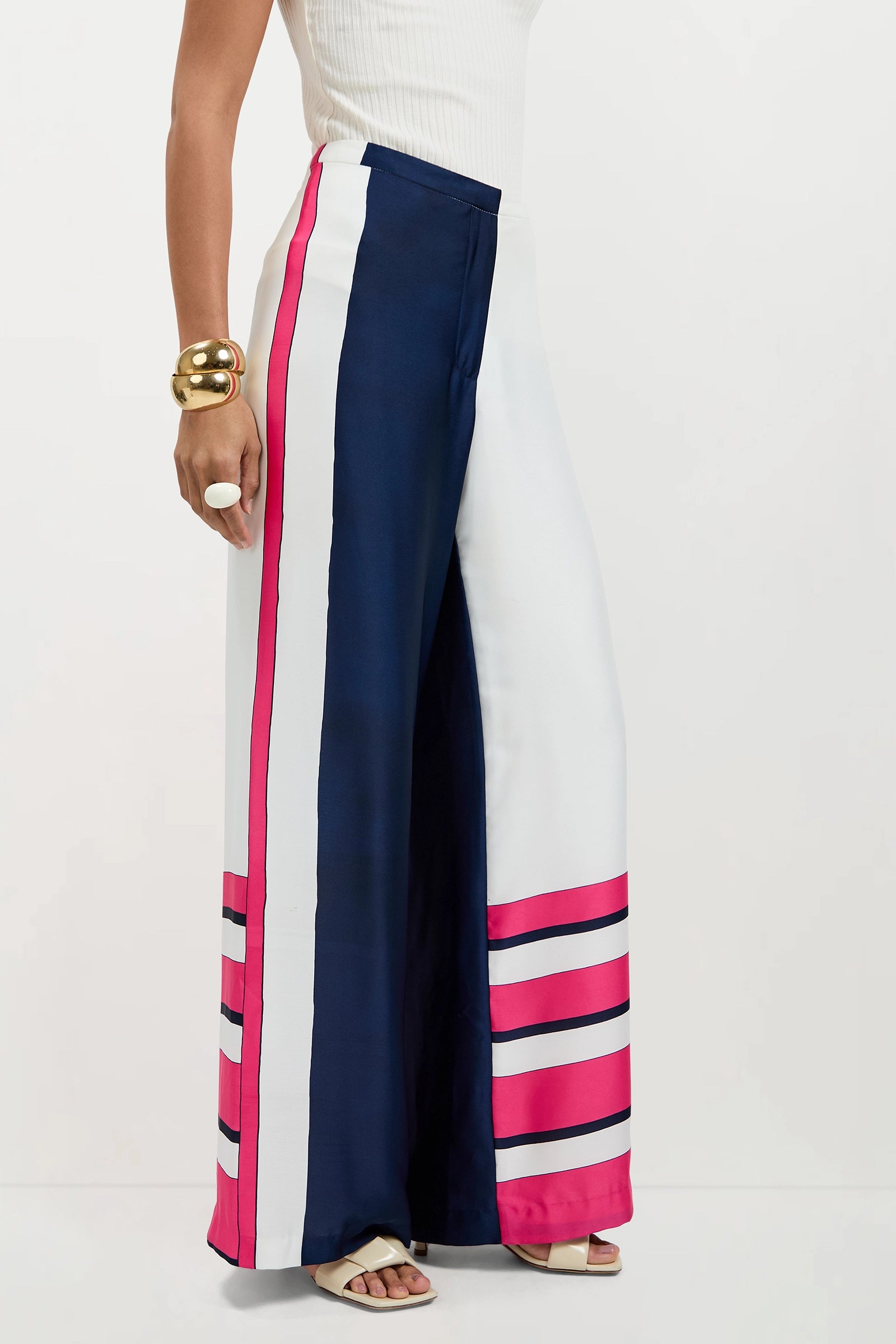 A person wears Lanay Linee Wide-Leg Pants with vertical navy, white, and pink stripes and horizontal pink stripes near the hem, styled with a sleeveless white top, chunky gold bracelet, and white sandals.