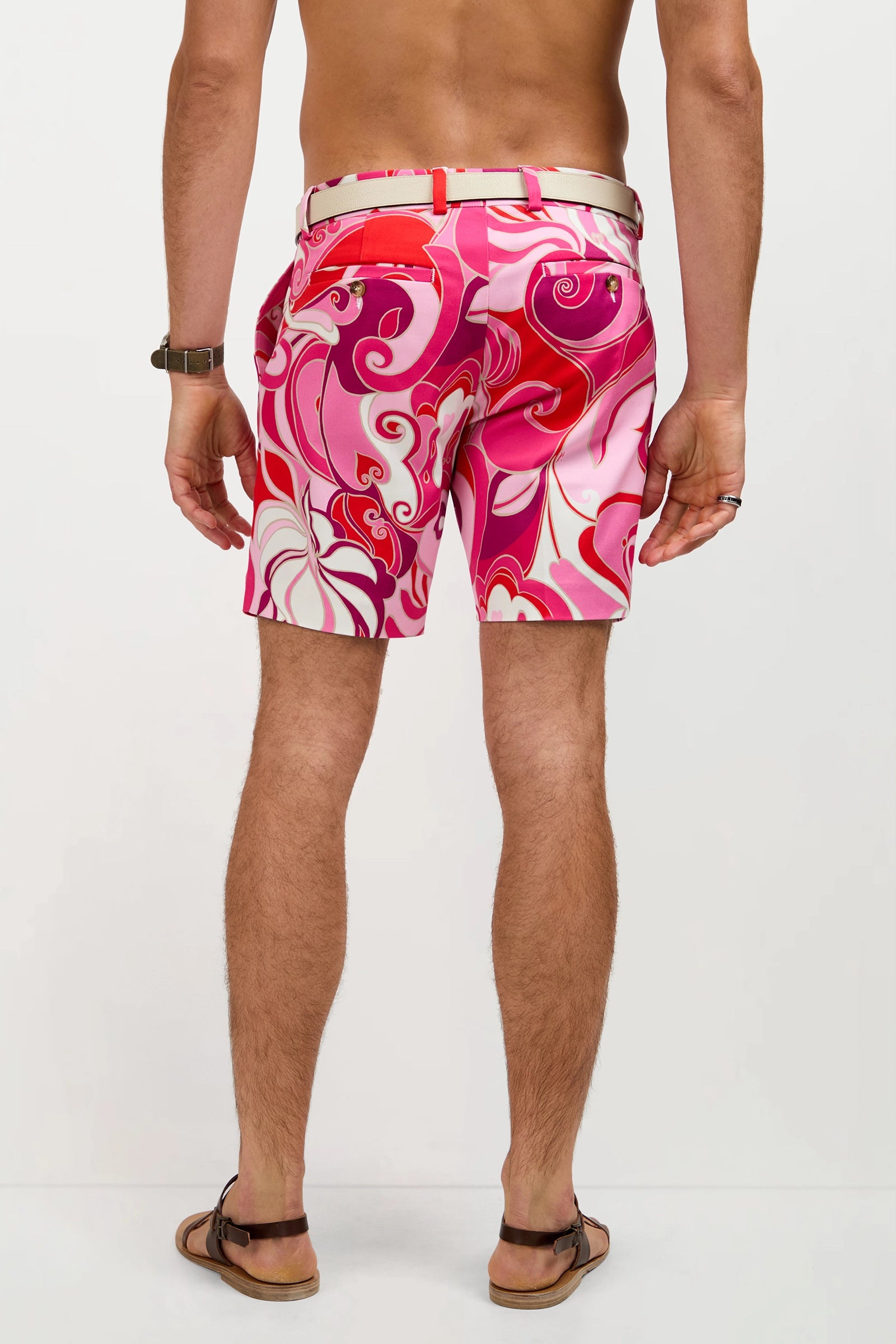 A man, shown from the waist down against a plain background, wears the Lawrence Short - Darsena with a vibrant pink, red, and white pattern, along with a watch, bracelet, ring, and brown sandals.