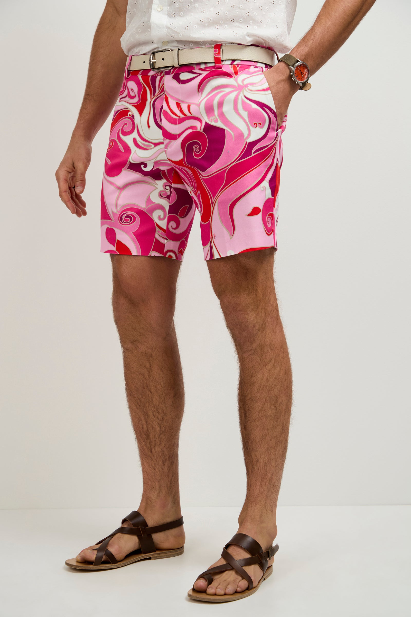 A person wearing the Lawrence Short - Darsena, featuring pink and red swirls, paired with a white shirt, beige belt, brown sandals, and a wristwatch, stands against a plain light background.