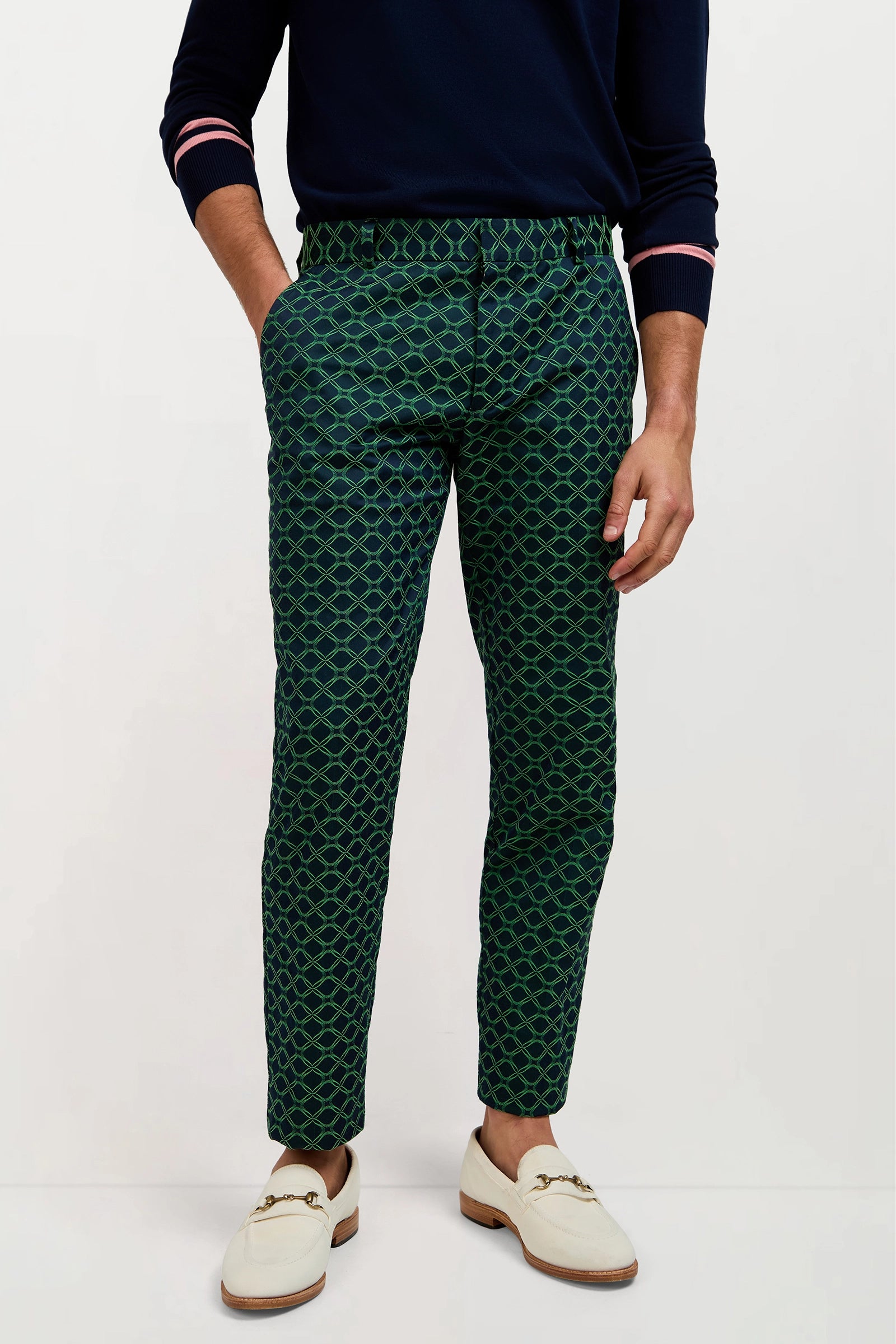 A man, head not shown, stands with hands in pockets against a white background wearing the Clyde Slim Trouser in Clover Geo Jacquard, a navy sweater with pink and white striped sleeves, and white loafers.
