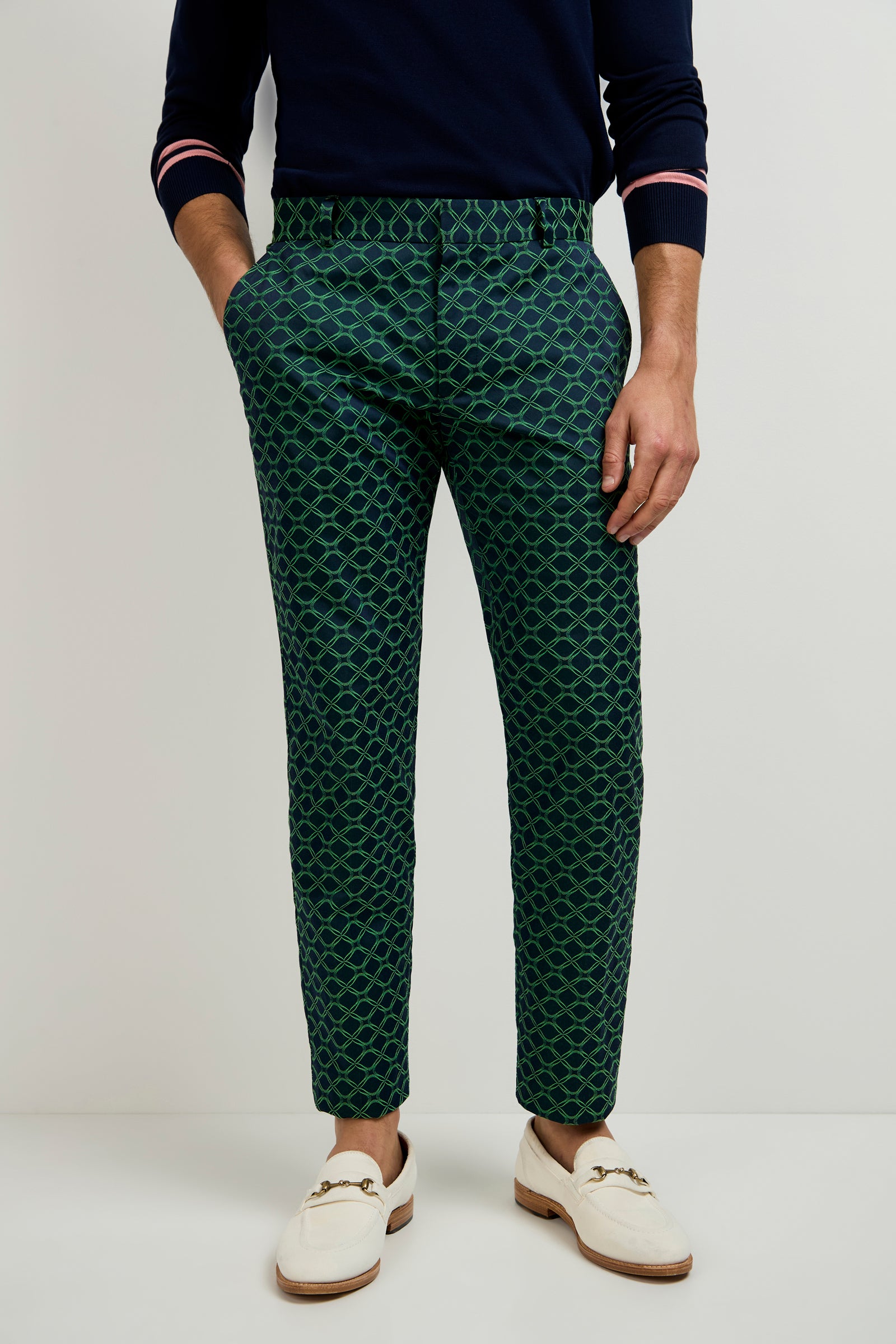 A person wears the Clyde Slim Trouser in Clover Geo Jacquard, paired with a navy sweater with pink-striped cuffs and white loafers. They stand against a plain light background, showing only the upper body and feet.