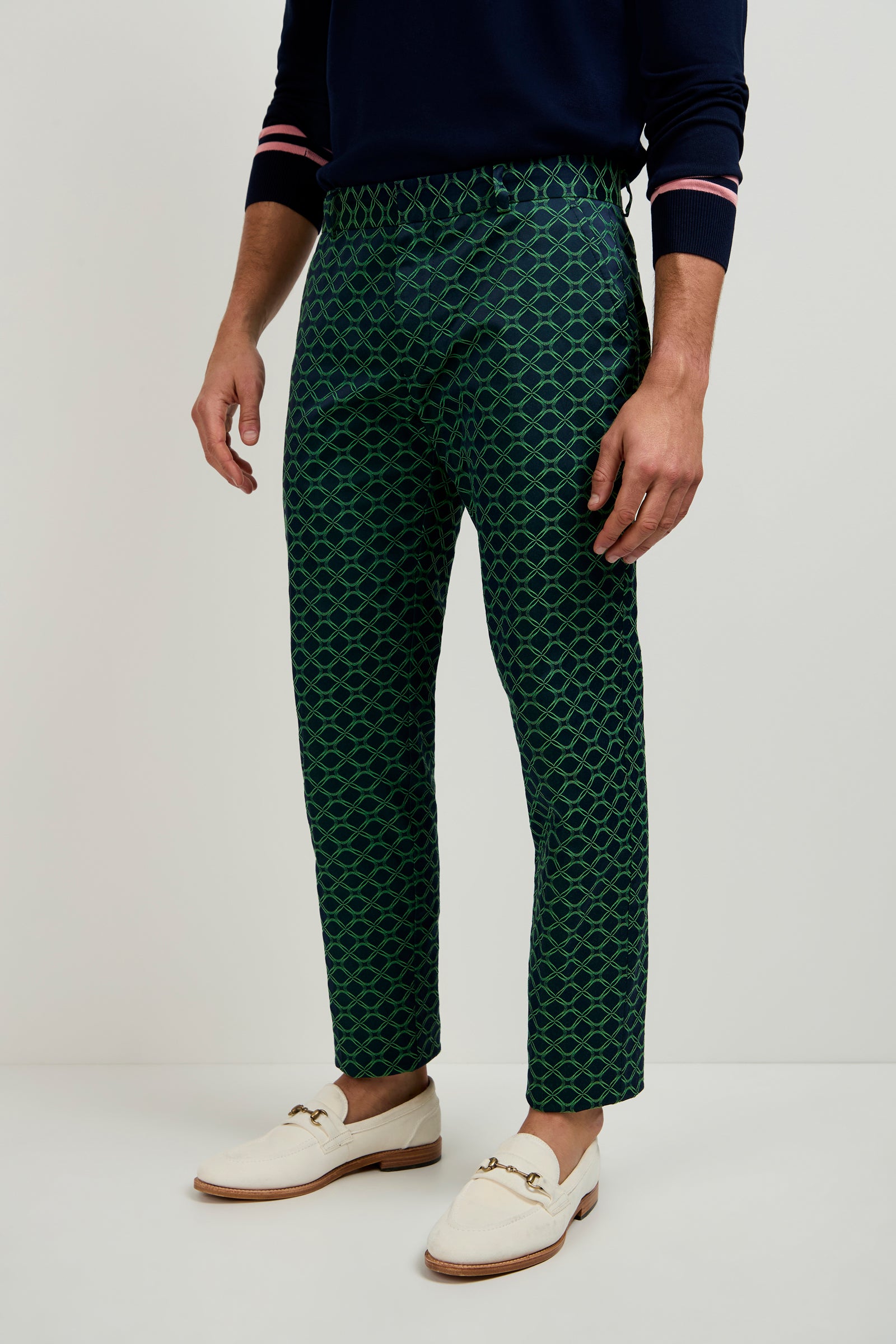 A person in the Clyde Slim Trouser - Clover Geo Jacquard, a navy sweater with pink-striped cuffs, and white loafers stands against a plain light background.