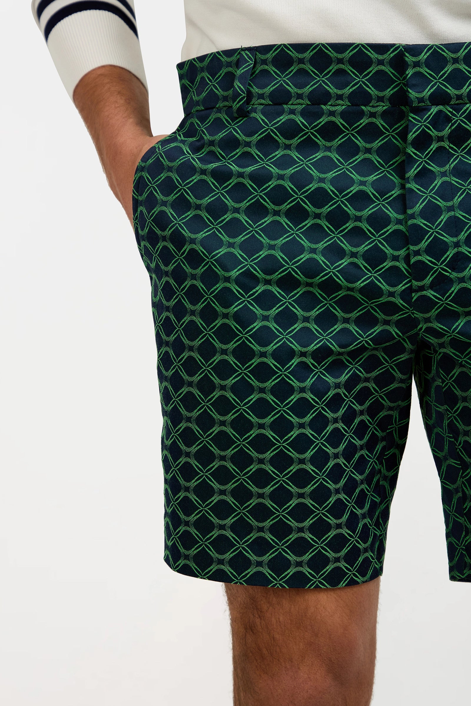 A person wears the Lawrence Short in Clover Geo Jacquard, featuring a dark green geometric pattern, paired with a white sweater with navy sleeve stripes. One hand is in the pocket. The image captures from waist to mid-thigh.