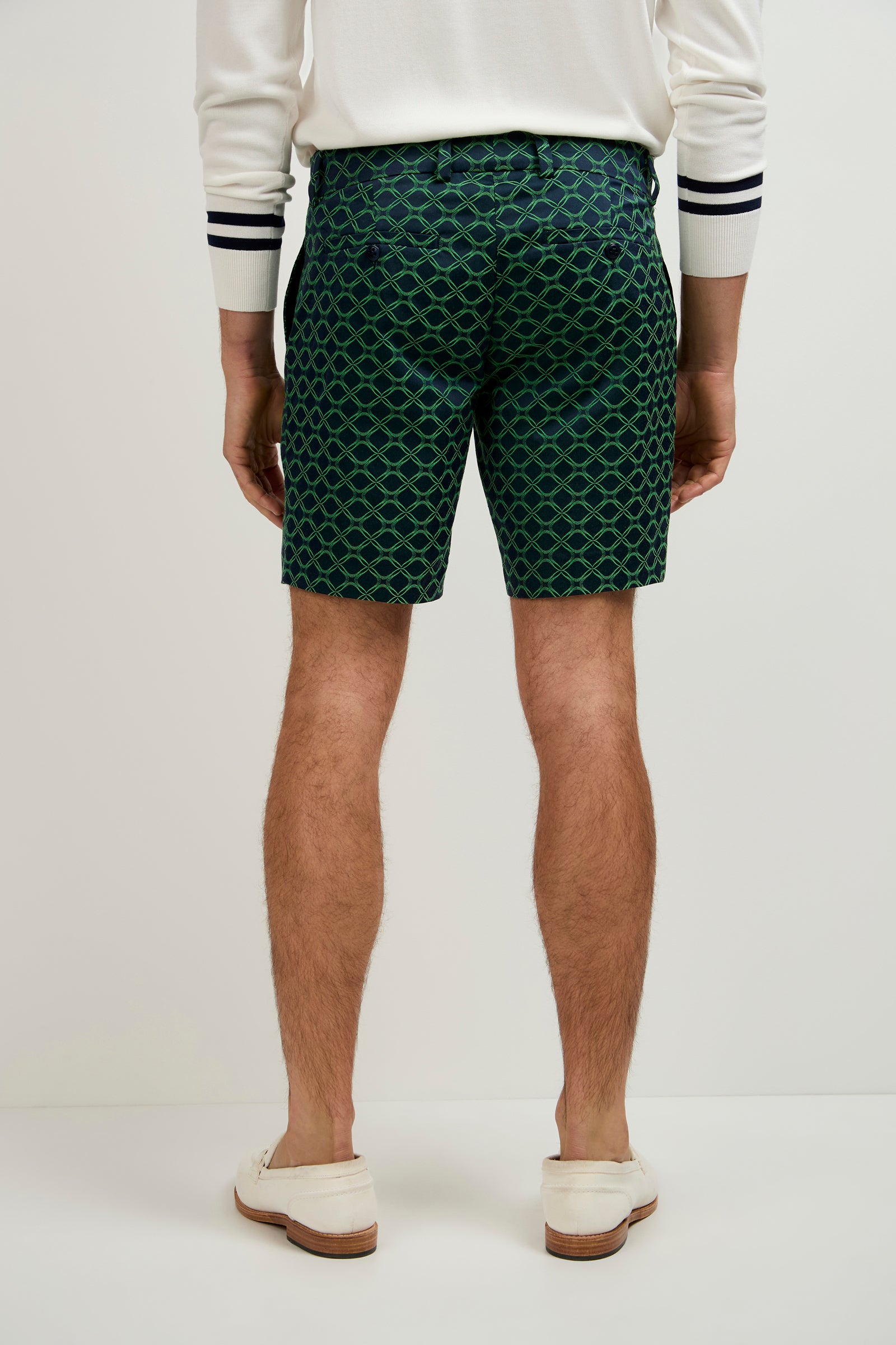 A person stands with their back to the camera, wearing a white long-sleeve shirt with black striped cuffs, Lawrence Short - Clover Geo Jacquard in green and blue patterns, and white loafers.