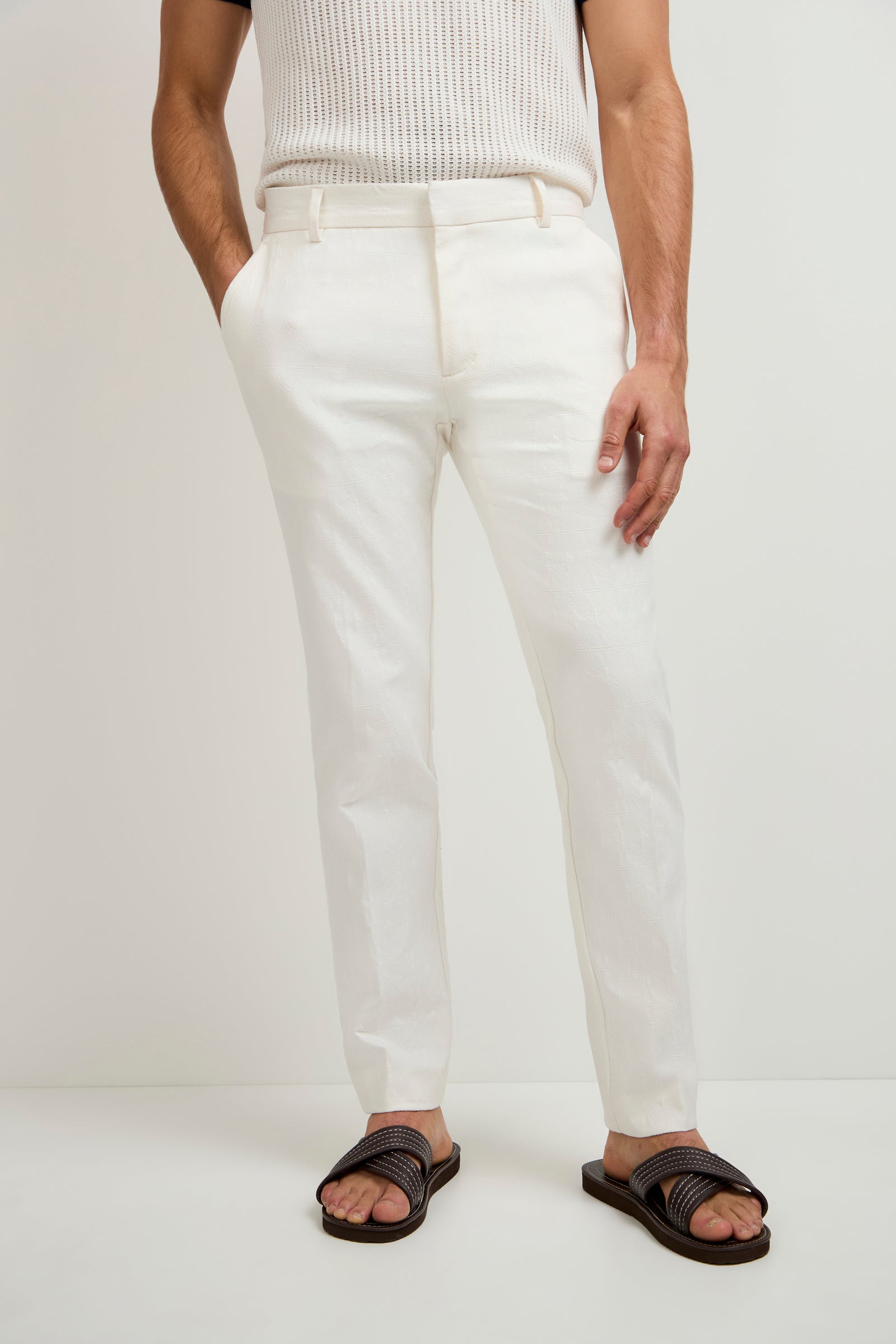 A person wears the Clyde Slim Trouser in Tegola Textured Jacquard, paired with a sleeveless ribbed white top and dark brown sandals, standing against a plain white background with hands in pockets.