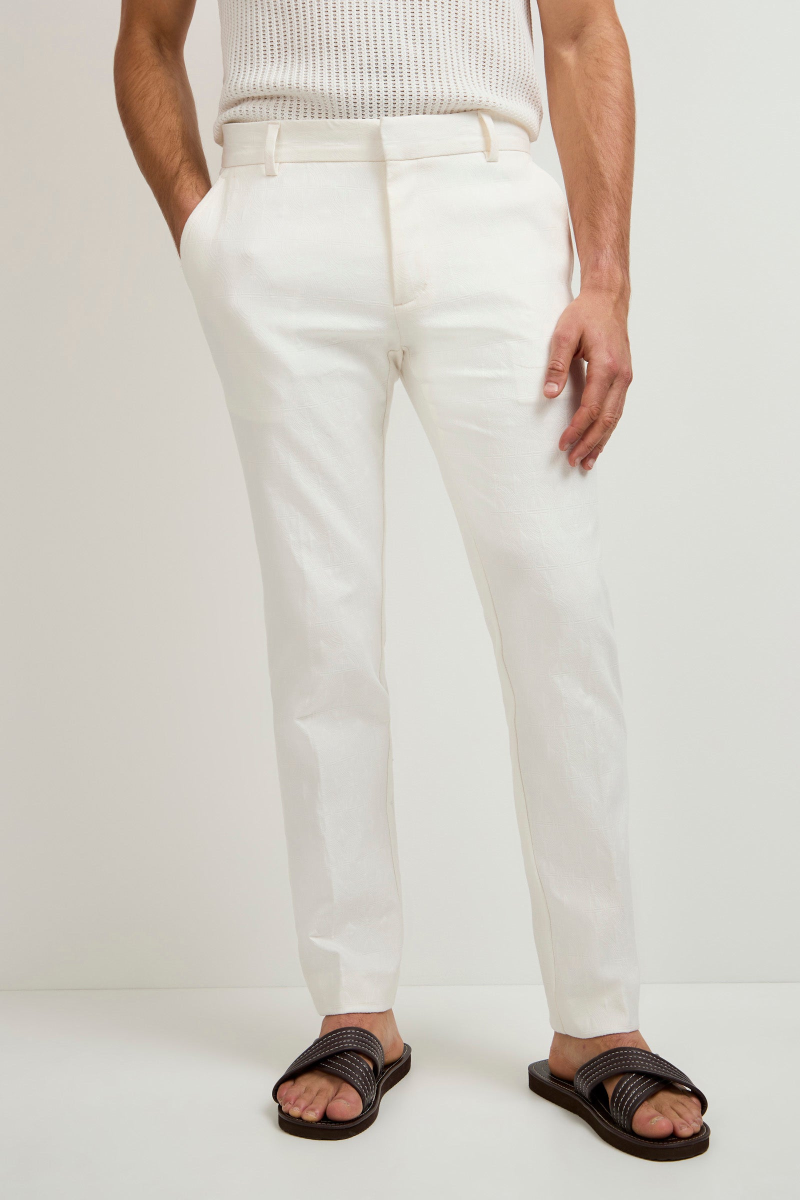 A person wearing the Clyde Slim Trouser - Tegola Textured Jacquard, a white sleeveless knit top, and brown sandals stands against a plain white background with hands in pockets.