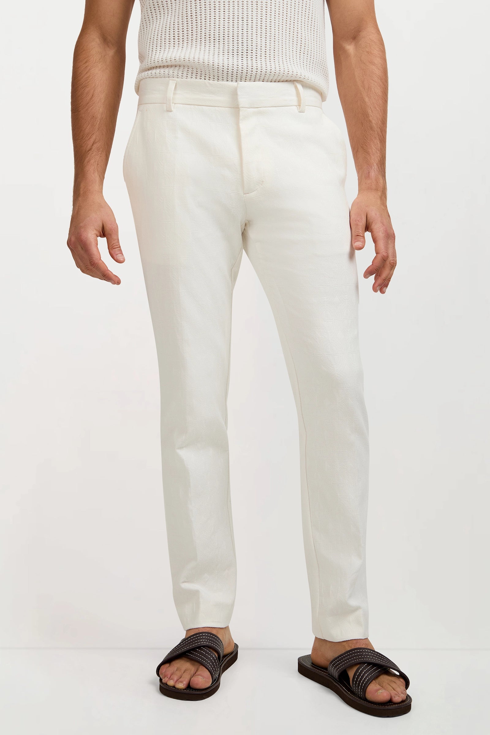 A person wearing the Clyde Slim Trouser - Tegola Textured Jacquard stands against a plain light background, paired with a white sleeveless textured top and dark woven sandals. Only their body from the shoulders down is visible.