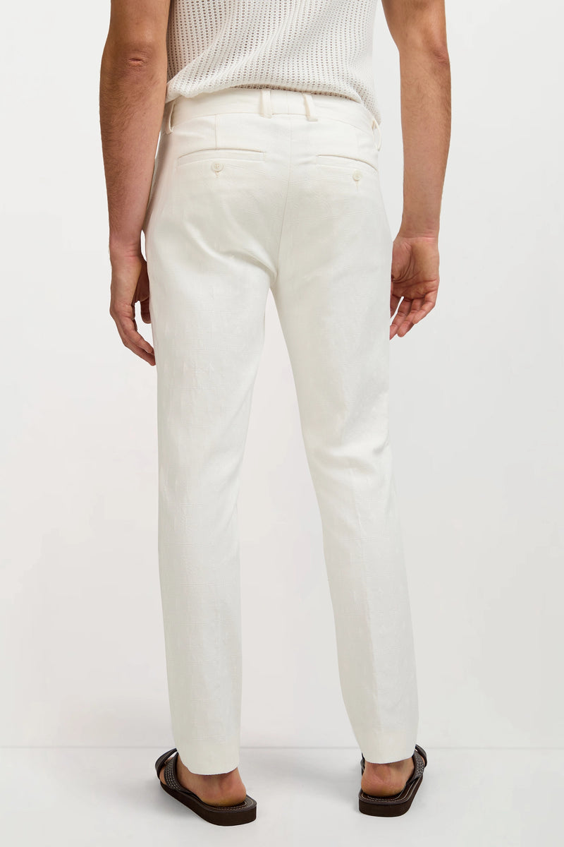 A man faces away from the camera, wearing Clyde Slim Trouser - Tegola Textured Jacquard, a white sleeveless knit top, and brown sandals against a plain light background.