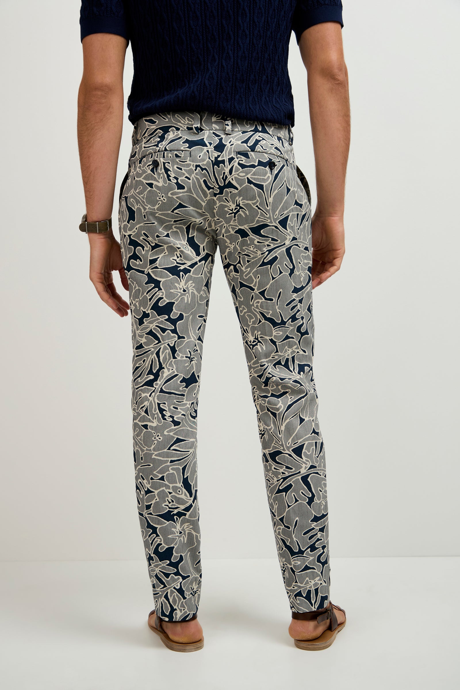 A person standing barefoot facing away, wearing a navy short-sleeve shirt and the Clyde Slim Trouser - Artsy Floral Jacquard, featuring a large beige and dark blue floral pattern. The background is plain white.