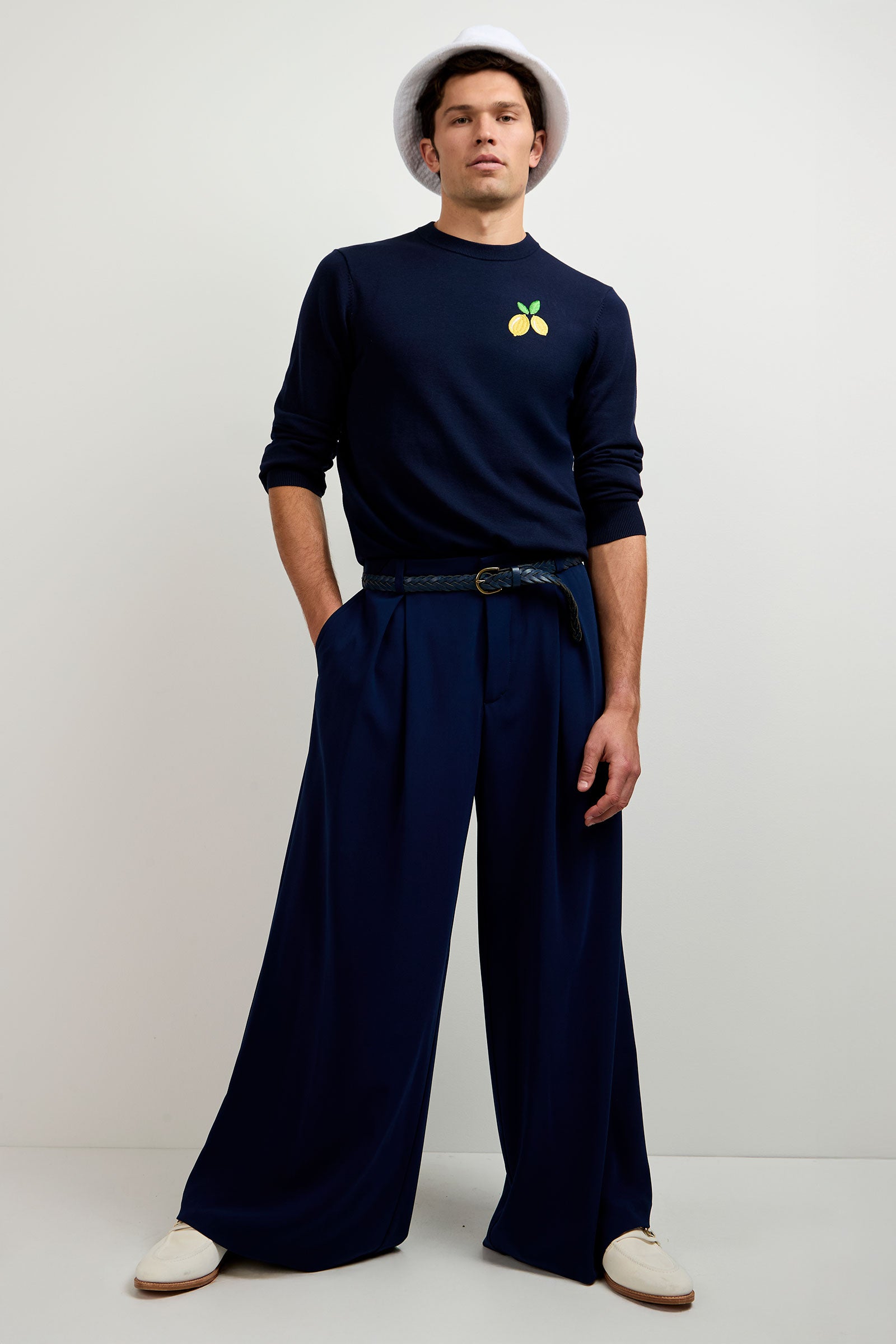 A man wearing a white hat, navy sweater with a lemon graphic, Clyde Wide-Leg Trouser in Ink, navy belt, and white shoes stands against a plain white background with his hands in his pockets.