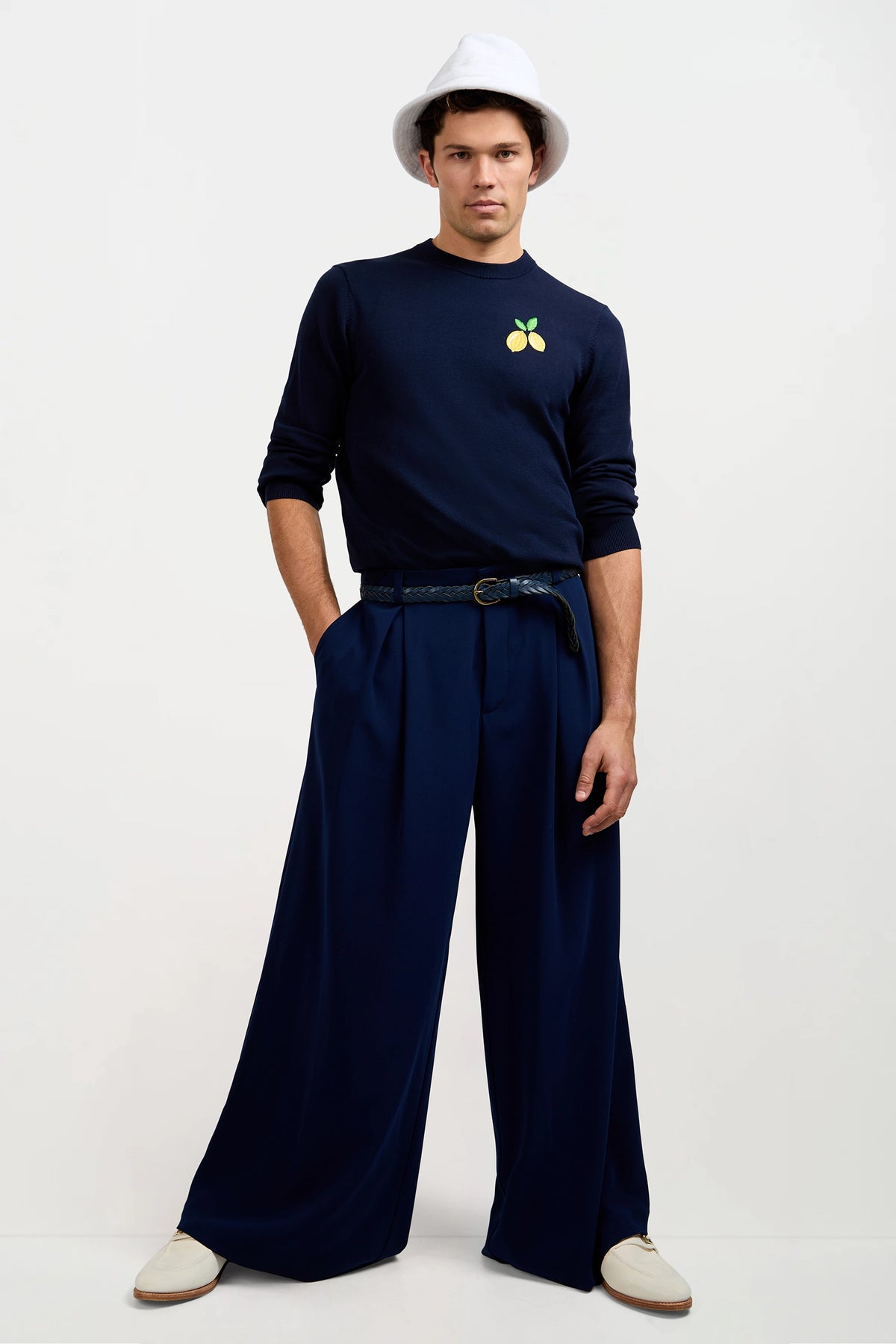 A man stands with hands in pockets, wearing a white bucket hat, a navy blue sweatshirt with a lemon patch, Clyde Wide-Leg Trouser in Ink, and white shoes against a plain background.