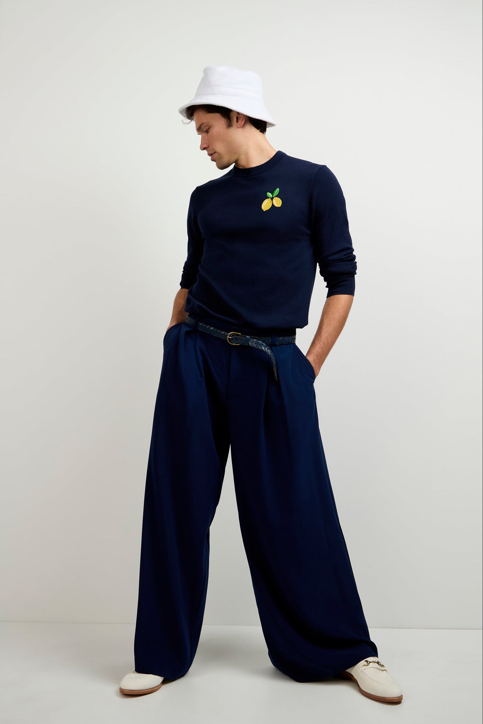 A man stands against a plain white background wearing ink-colored Clyde Wide-Leg Trousers, a navy sweater with a small fruit graphic, a white bucket hat, black belt, and white shoes.