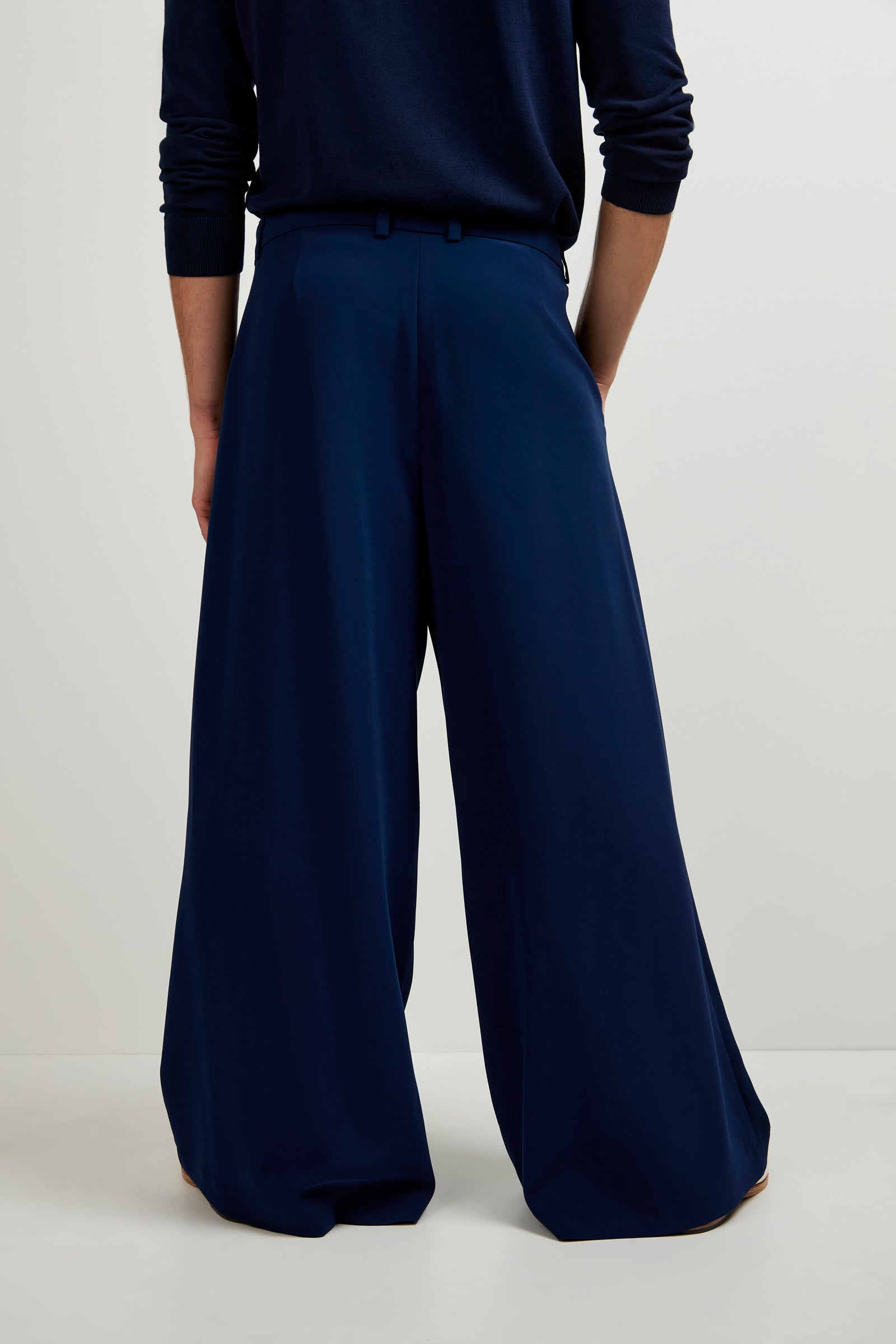 A person stands against a plain background, hands in pockets, wearing Clyde Wide-Leg Trousers in navy blue with a matching long-sleeved top. The head is not visible.