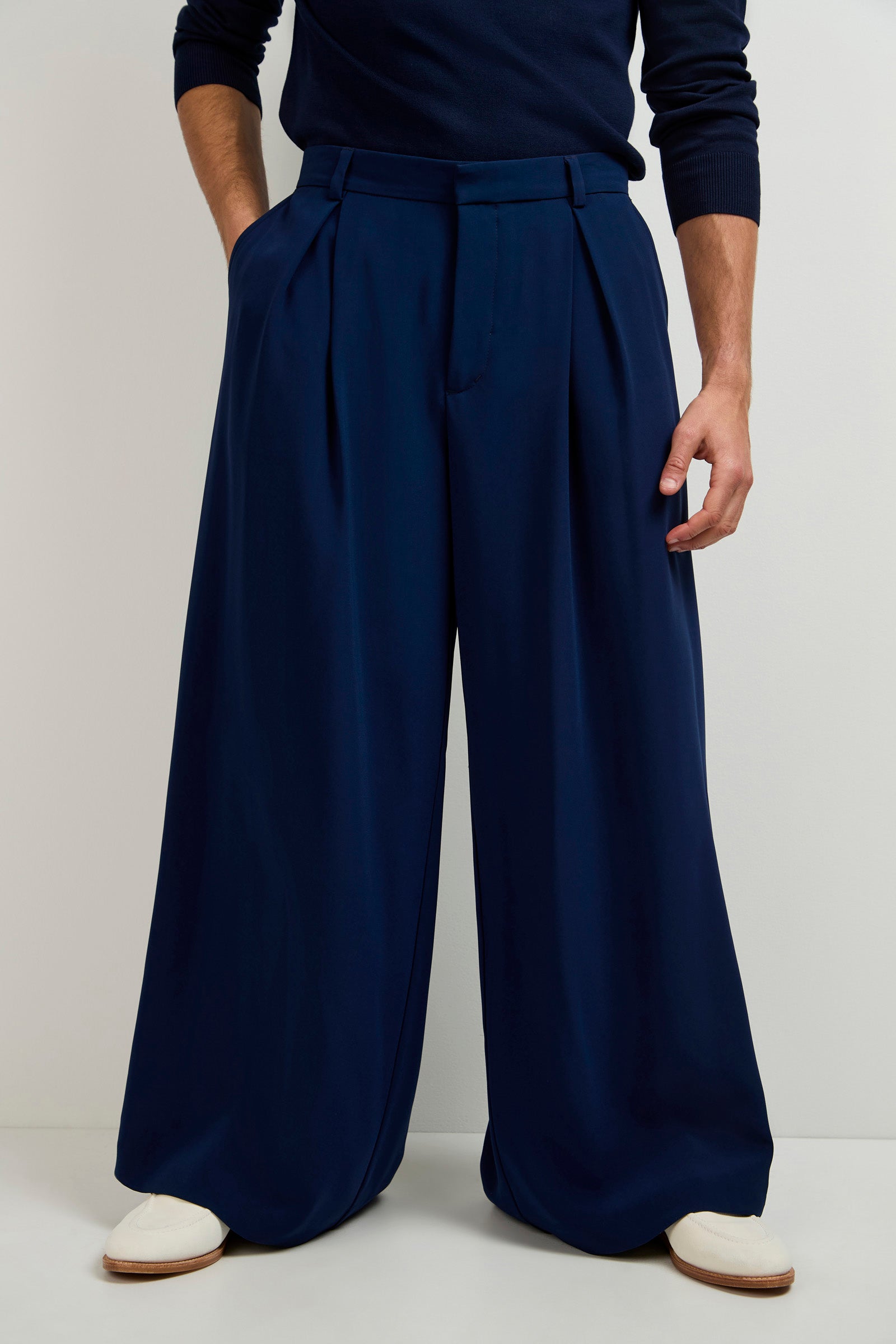 A person in the Clyde Wide-Leg Trouser in dark blue, featuring deep pleats, paired with a navy sweater and white shoes, stands against a plain light background. The image is cropped above the shoulders.