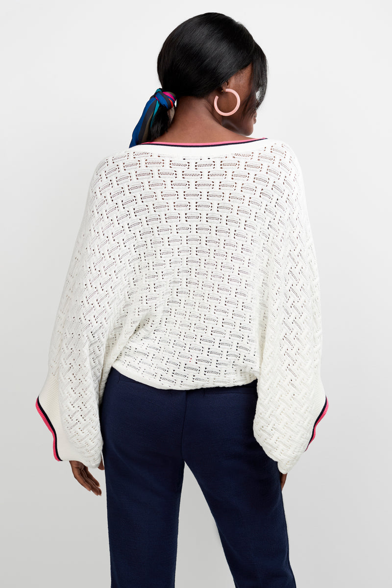 A person with dark hair tied with a colorful scarf is seen from behind wearing the Horizon Boatneck Pullover—a loose white knit sweater with wide sleeves—paired with navy blue pants against a plain white background.