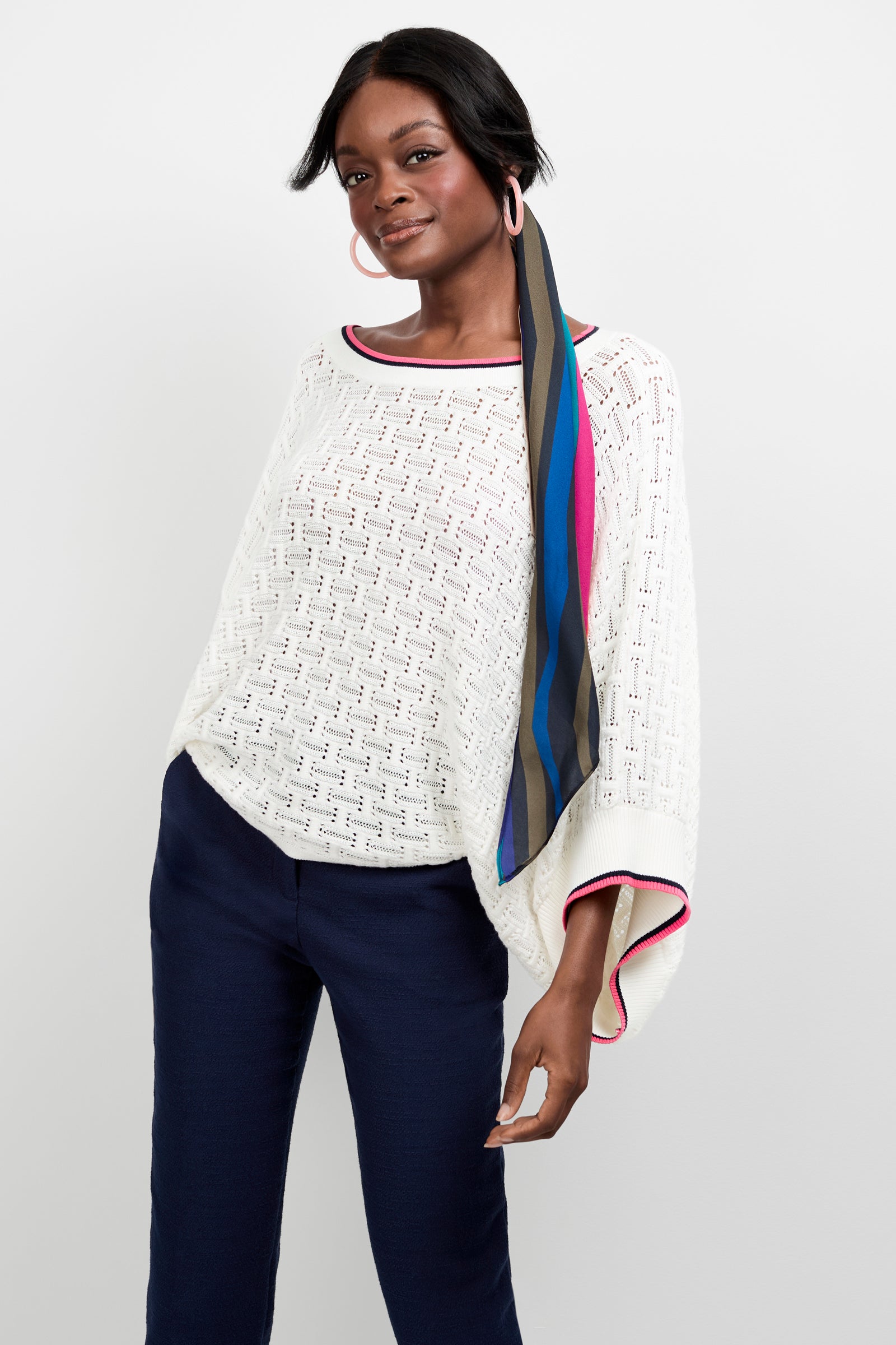 A woman wears the Horizon Boatneck Pullover—a white, loose-knit sweater with pink trim—paired with navy blue pants, large pink hoop earrings, and a colorful striped hair scarf, standing against a plain white background.