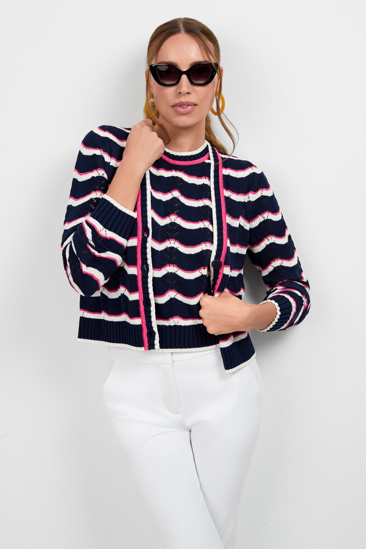 A woman stands against a plain white background wearing black sunglasses, white pants, and the Alessia Knit Cardigan in navy blue, white, and pink stripes—one hand touches her sunglasses, the other holds her cardigan.