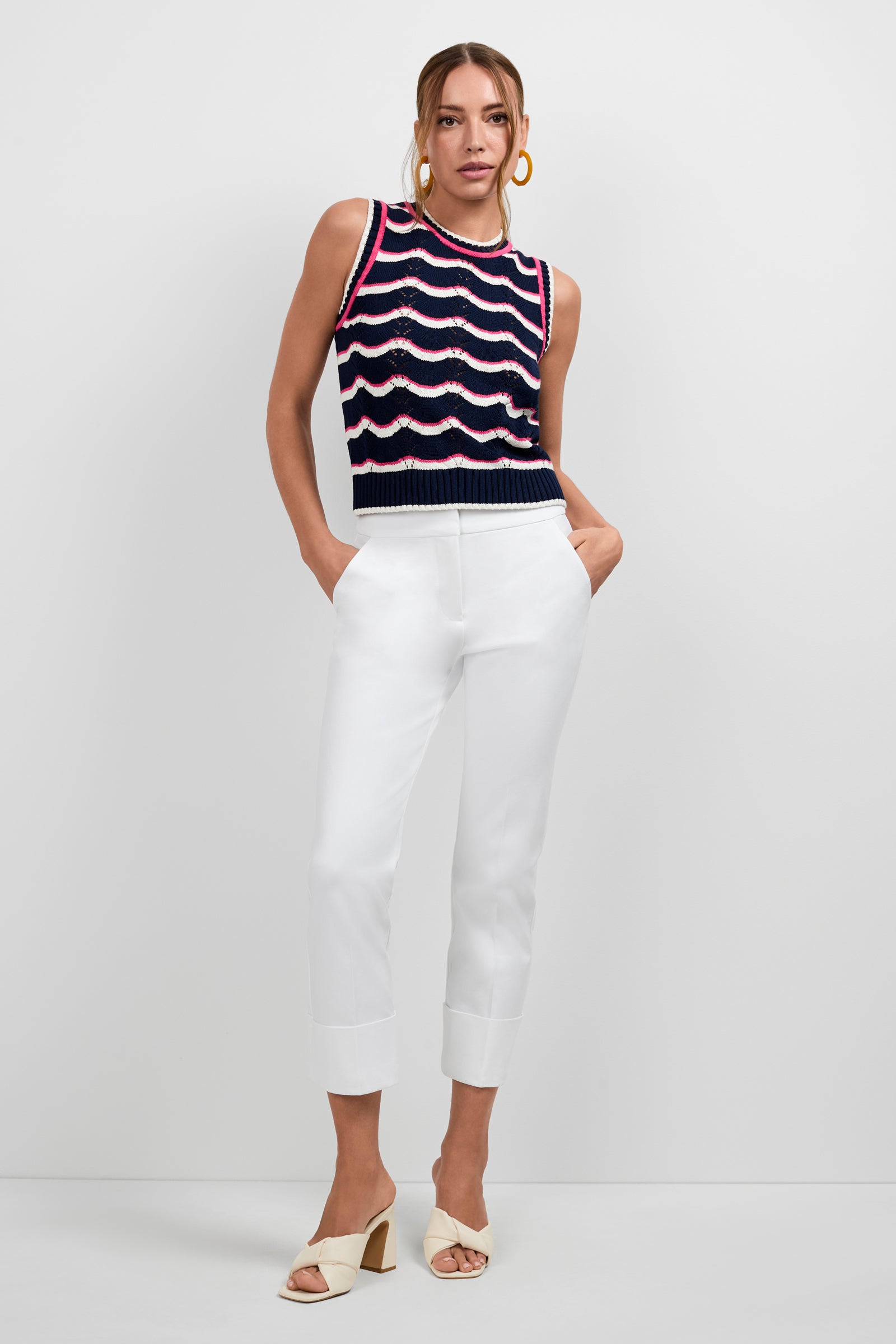 A woman stands against a white background wearing the Banshee Cropped Cuff Hem Pants, a sleeveless navy top with wavy red and white stripes, beige heeled sandals, and large hoop earrings. Her hands are in her pockets.